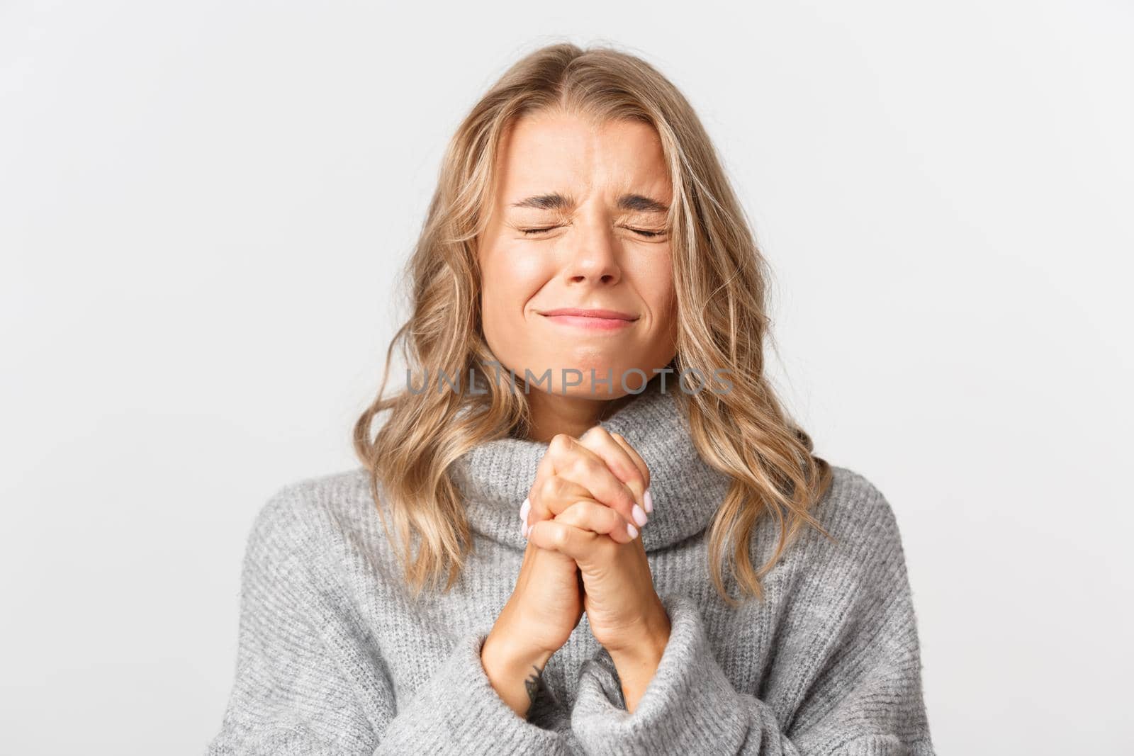 Close-up of hopeful blond girl waiting for chance, close eyes and clenching hands, pleading, standing over white background.