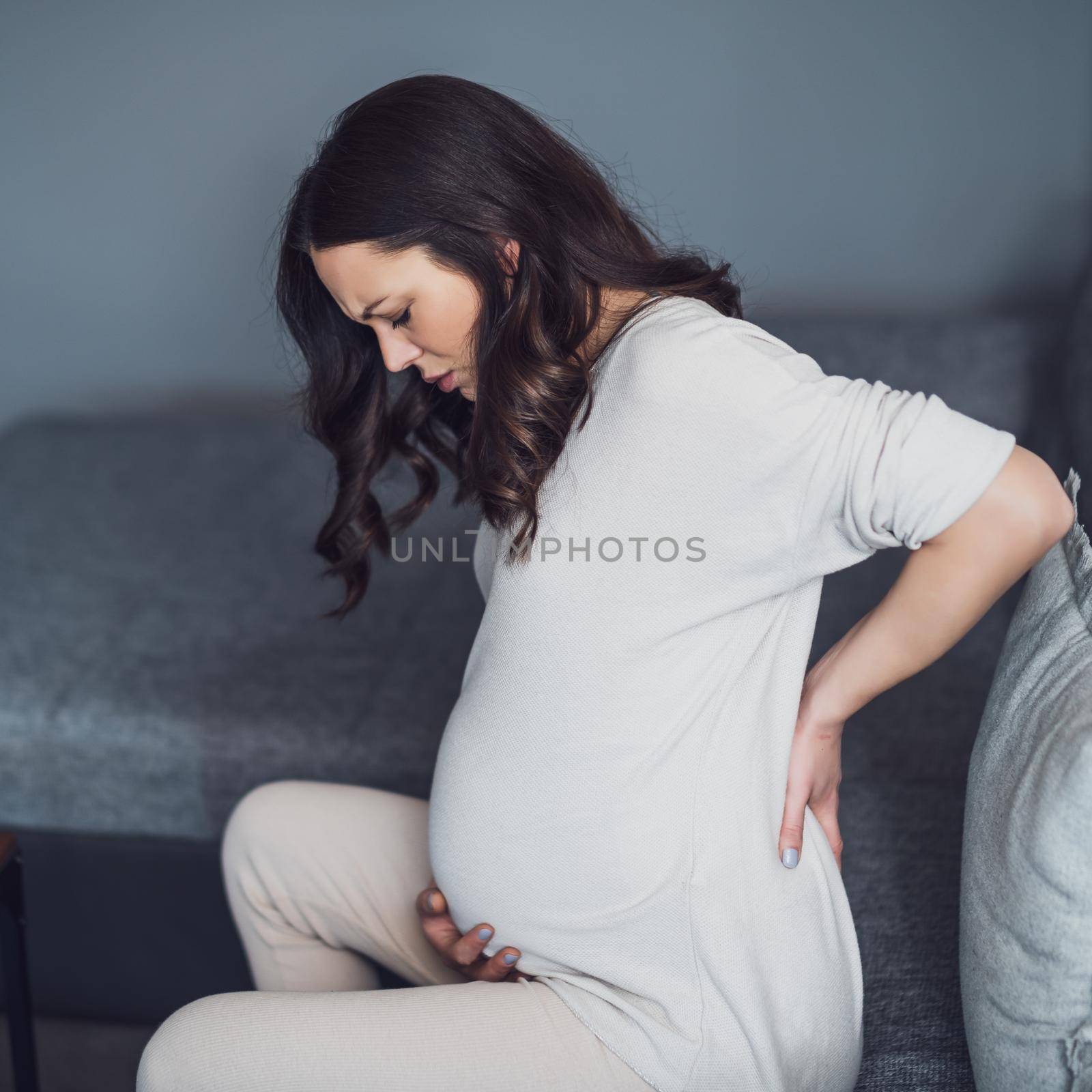 Pregnant woman is having pain in her back. She is sitting on bed at home.