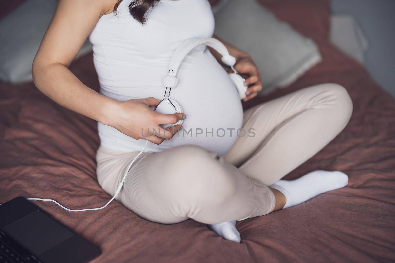 Close up of pregnant woman relaxing at home. She is sitting on bed in bedroom and playing music to her baby in stomach.