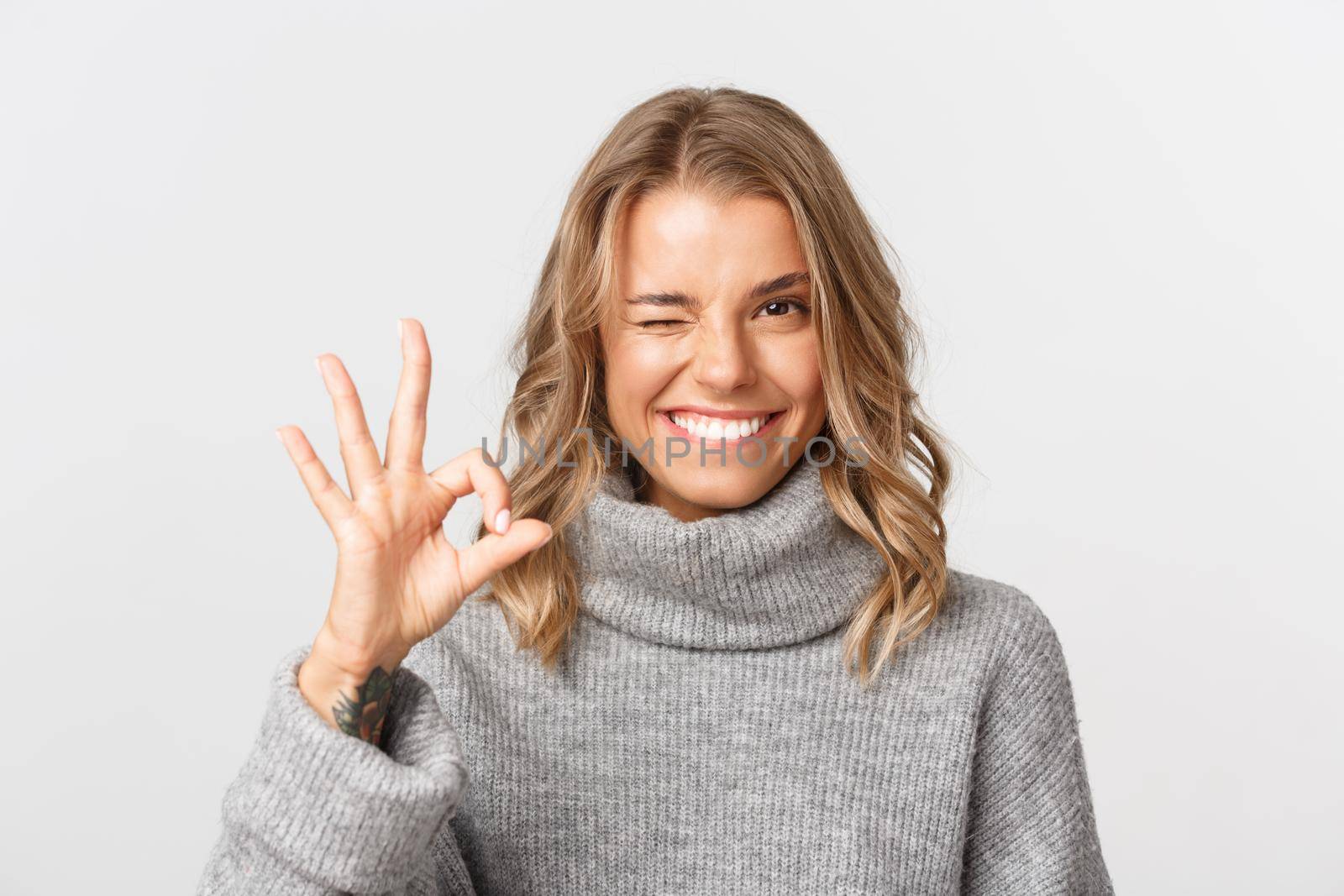 Close-up of confident pretty girl in grey sweater, showing okay sign and smiling, guarantee something, give approval, standing over white background.