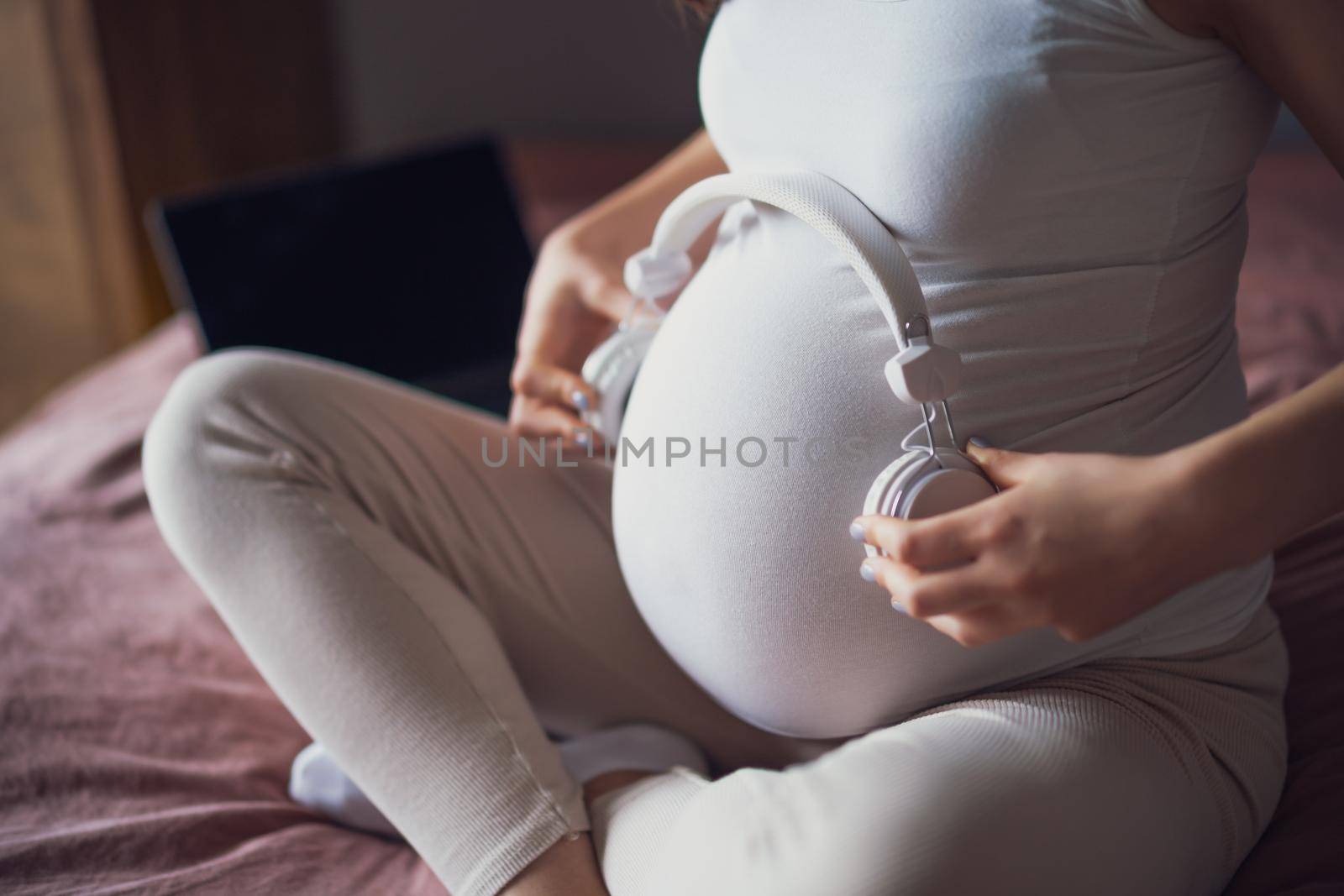 Close up of pregnant woman relaxing at home. She is sitting on bed in bedroom and playing music to her baby in stomach.