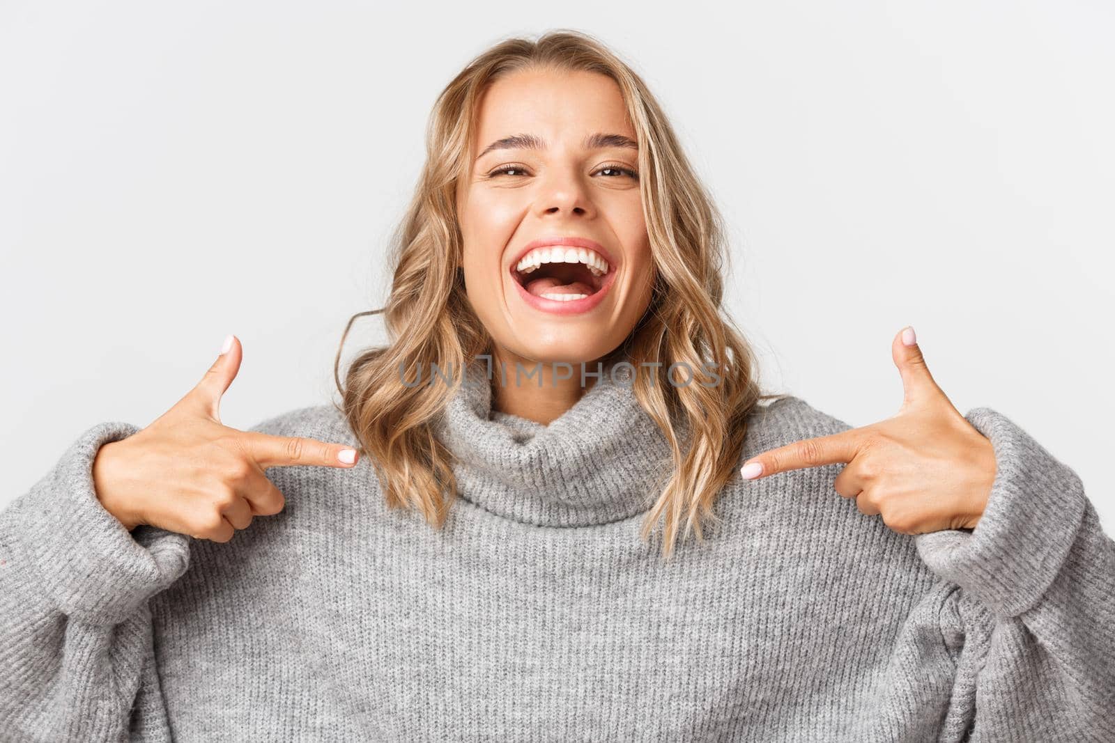 Close-up of happy attractive blond girl laughing, smiling and pointing fingers at your logo, standing over white background.