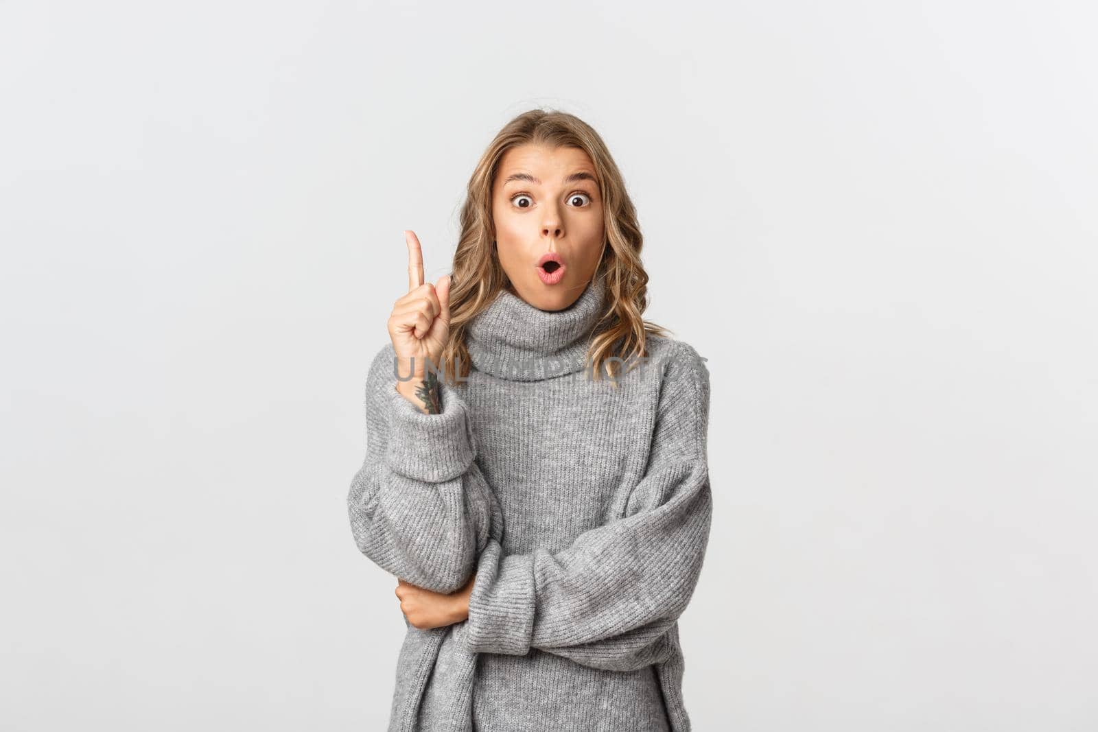 Close-up of thoughtful cute girl in grey sweater, having an idea, raising finger and say something, standing over white background.