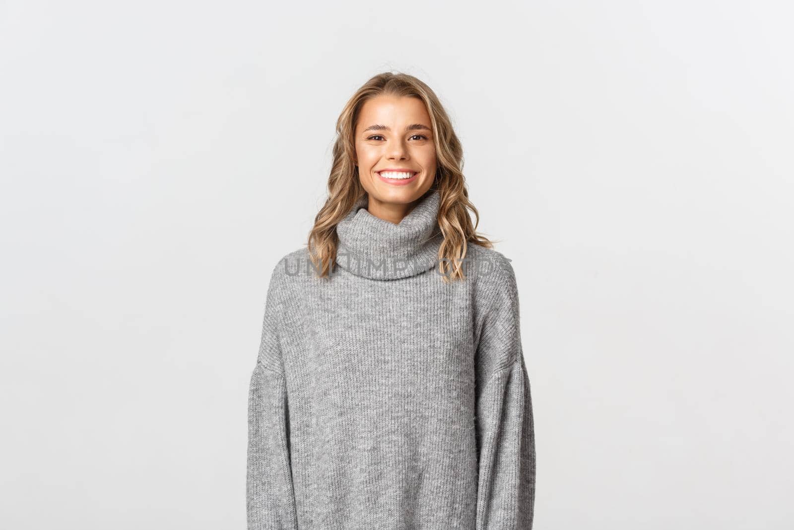 Image of beautiful happy woman in grey sweater, smiling and looking pleased, standing over white background.