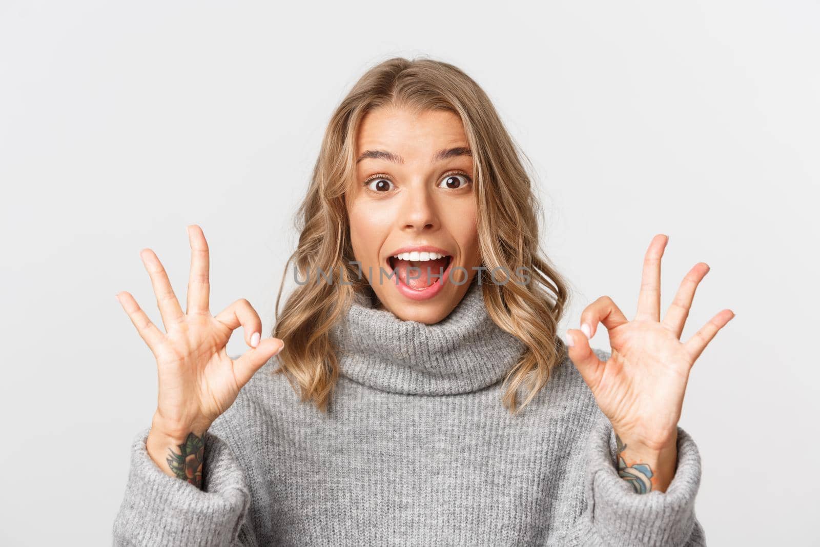 Close-up of attractive blond girl, showing okay signs and smiling excited, standing over white background by Benzoix