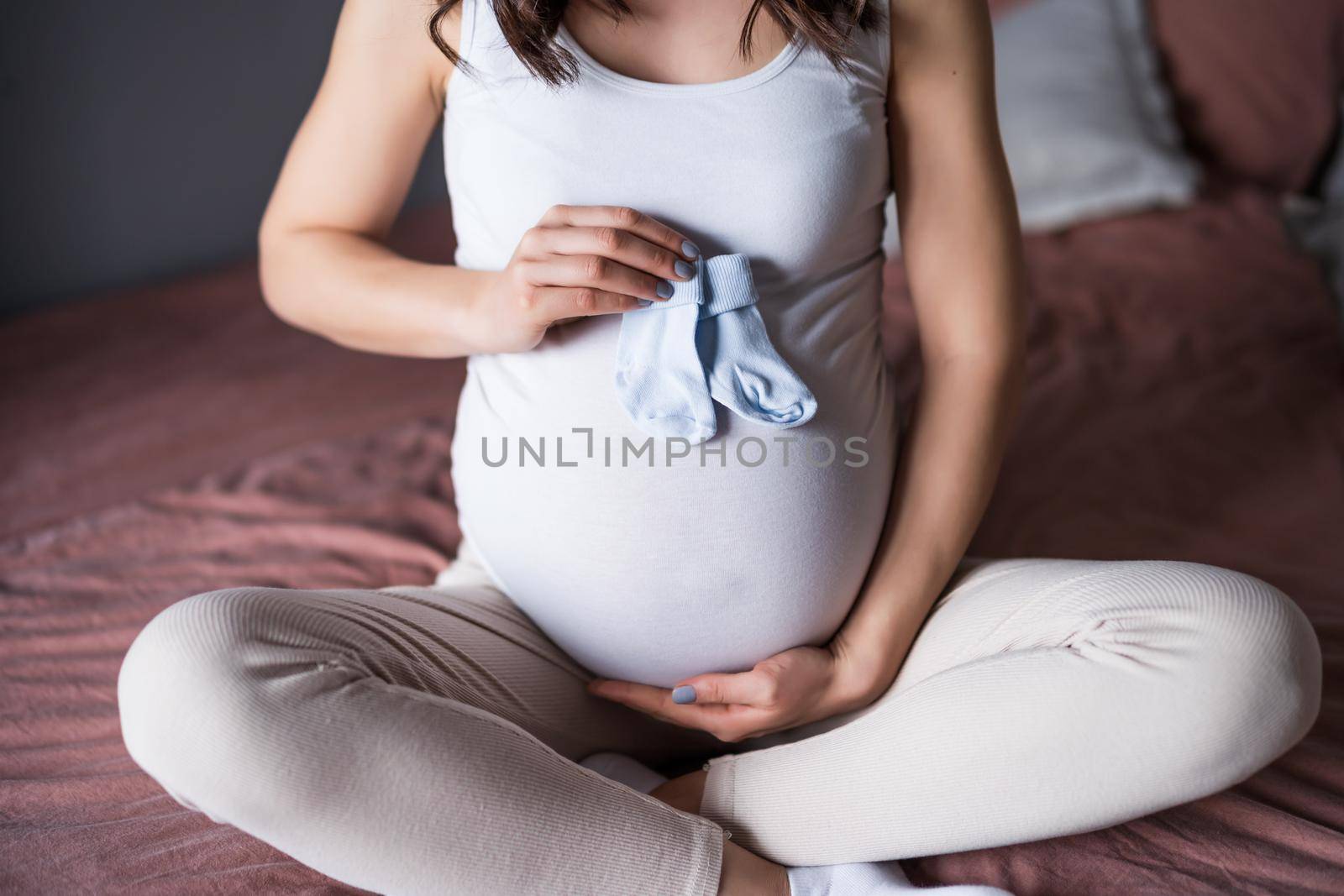 Pregnant woman relaxing at home. She is sitting on bed and holding baby socks.
