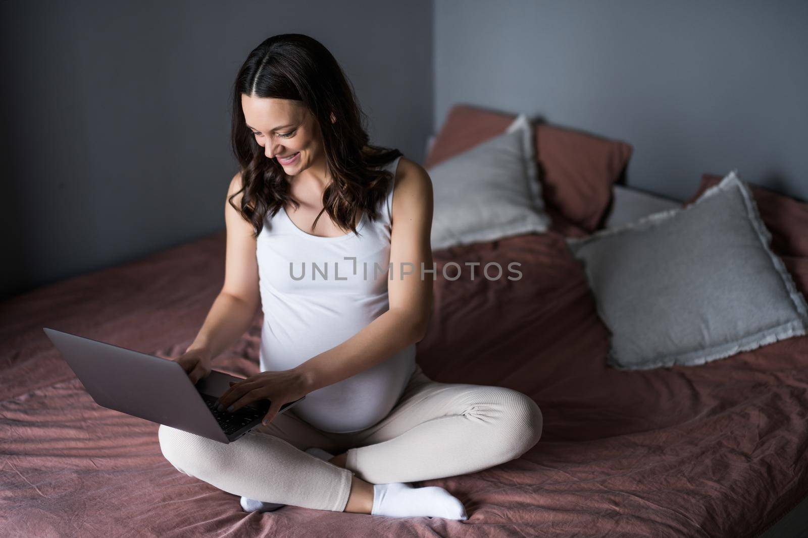 Happy pregnant woman relaxing at home. She is sitting on bed in bedroom and using laptop.