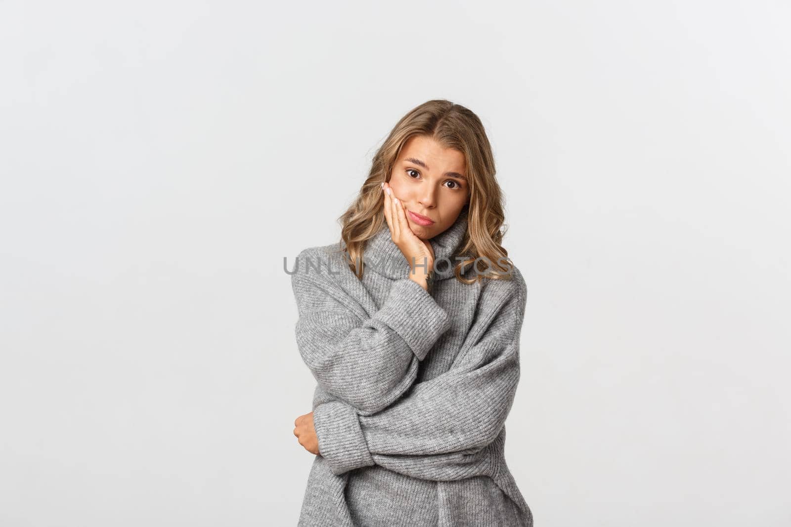 Portrait of sad beautiful female model in grey sweater, looking at camera with gloomy face, standing over white background by Benzoix