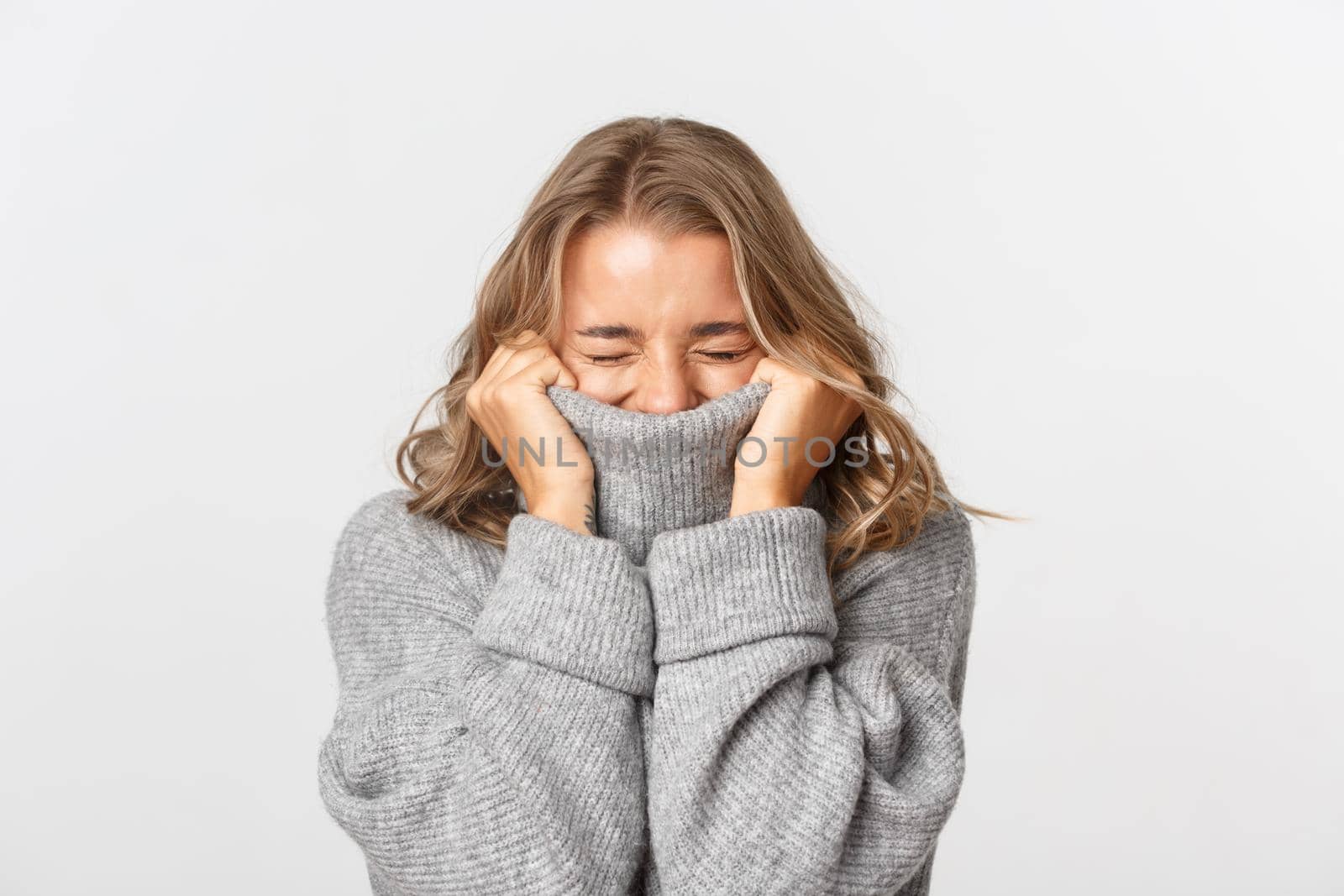 Close-up of happy blond girl, pulling sweater on face and smiling cheerful, standing over white background.