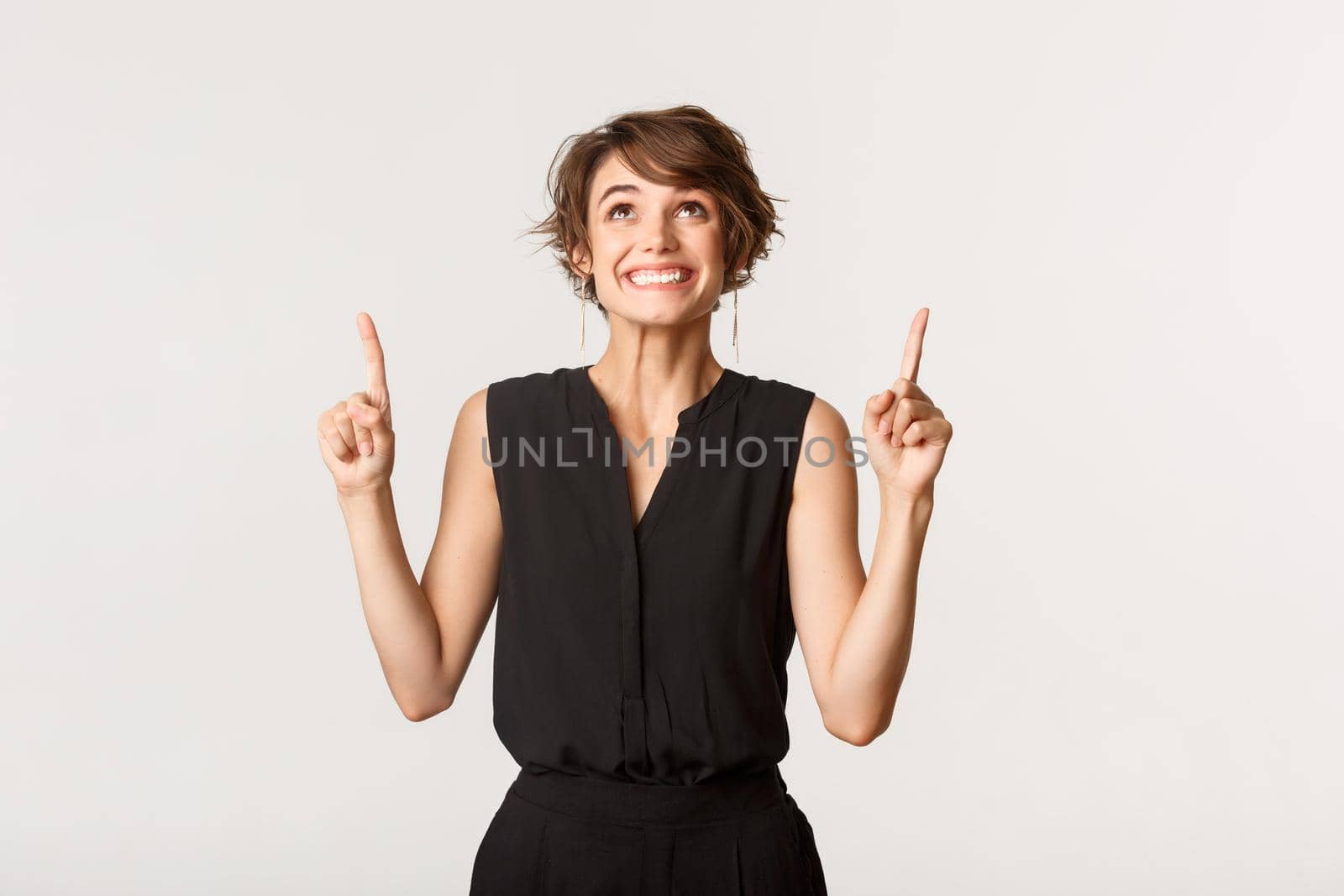 Excited beautiful young woman smiling and pointing fingers up with rejoice, standing white background.