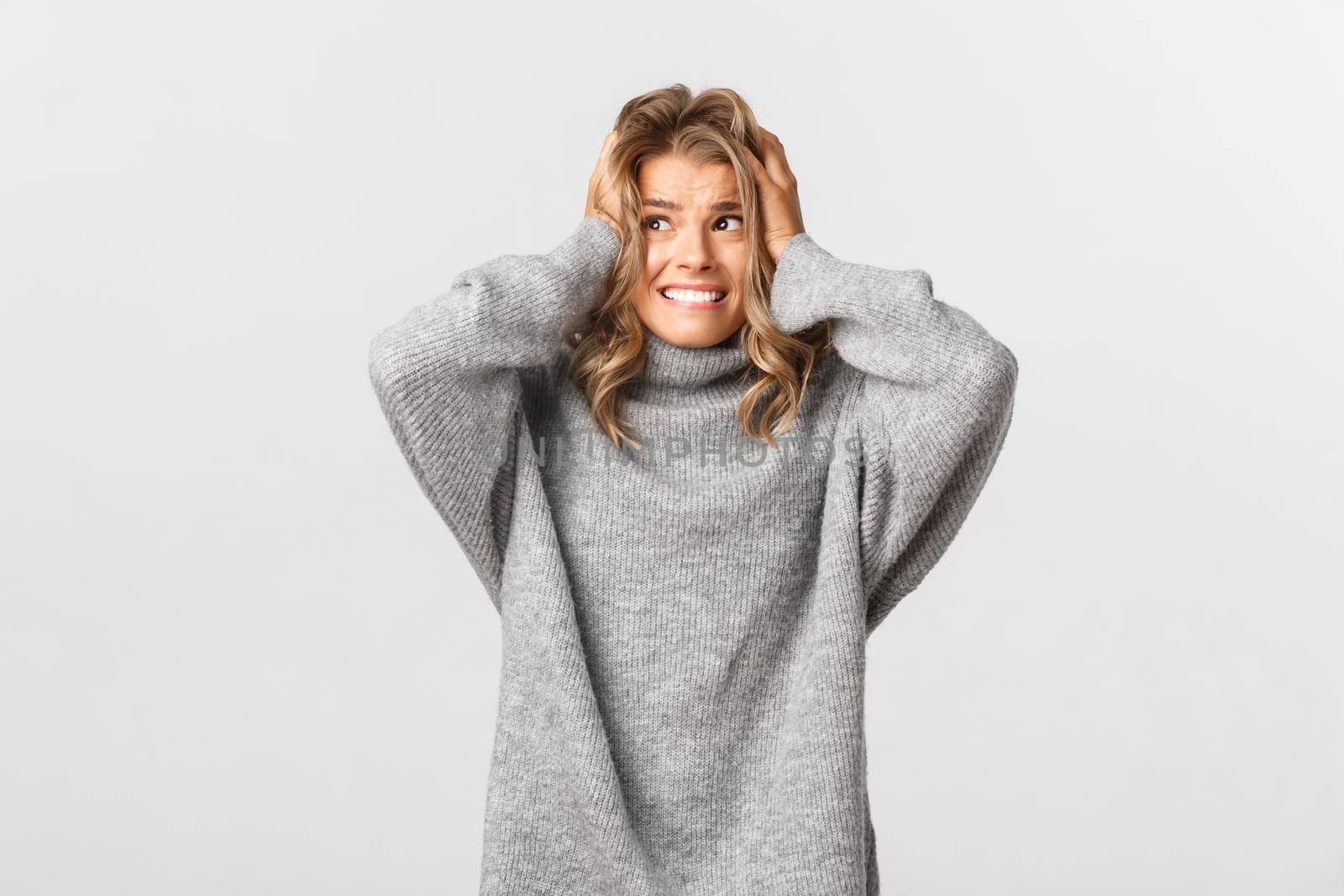 Portrait of blond girl in panic, looking left and holding hands on head, feeling alarmed and distressed, standing over white background.