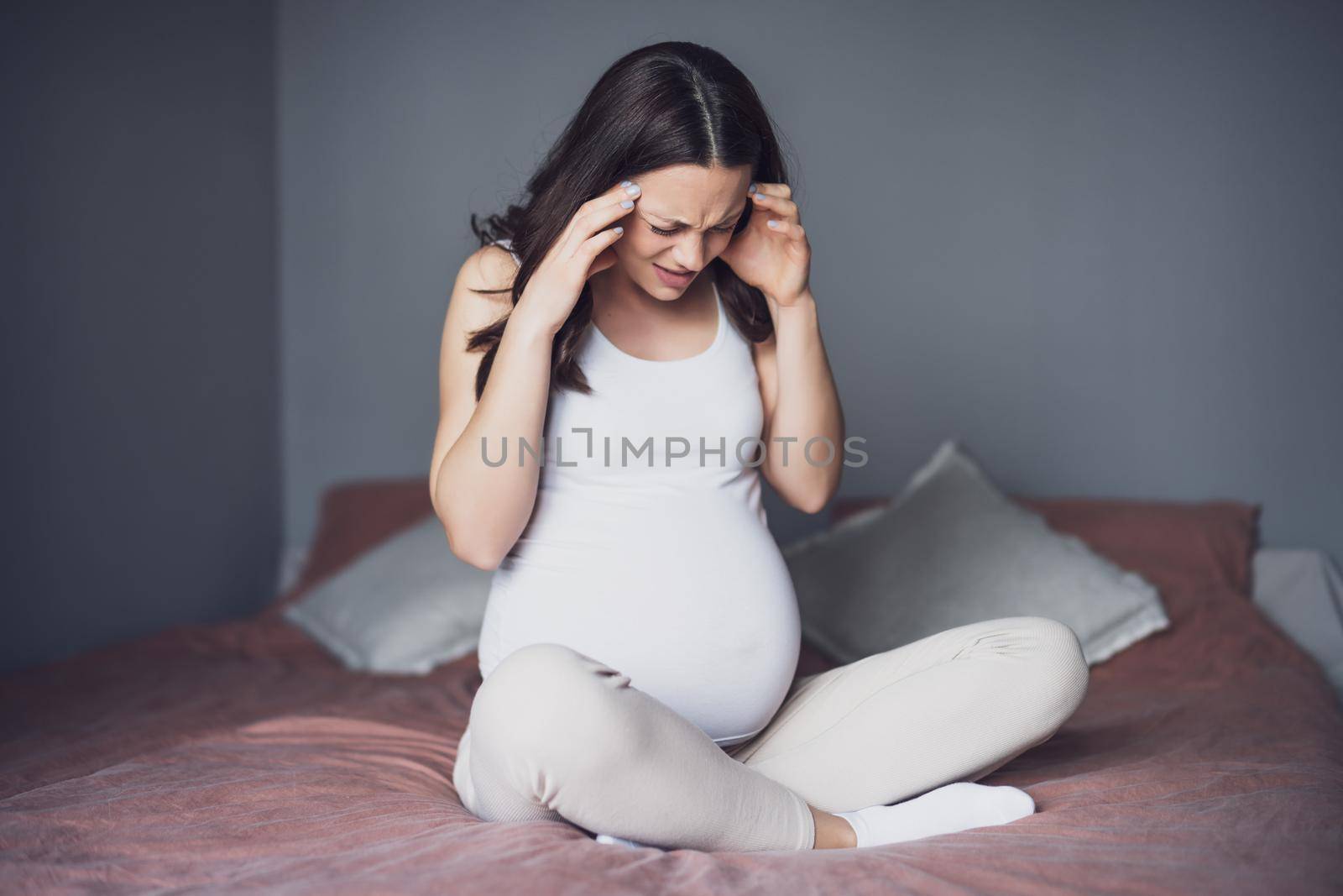 Pregnant woman is having headache. She is sitting on bed at home.