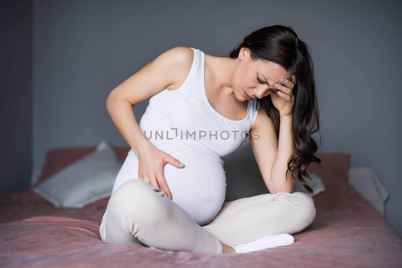 Pregnant woman is having pain in her stomach. She is sitting on bed at home.