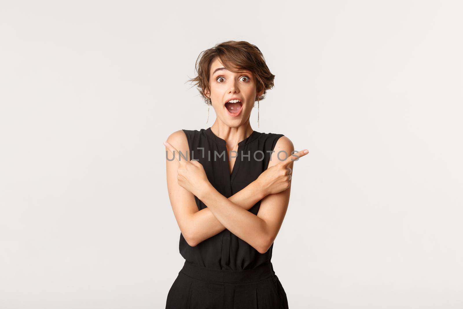 Excited young woman pointing fingers sideways at two products, making choice over white background.