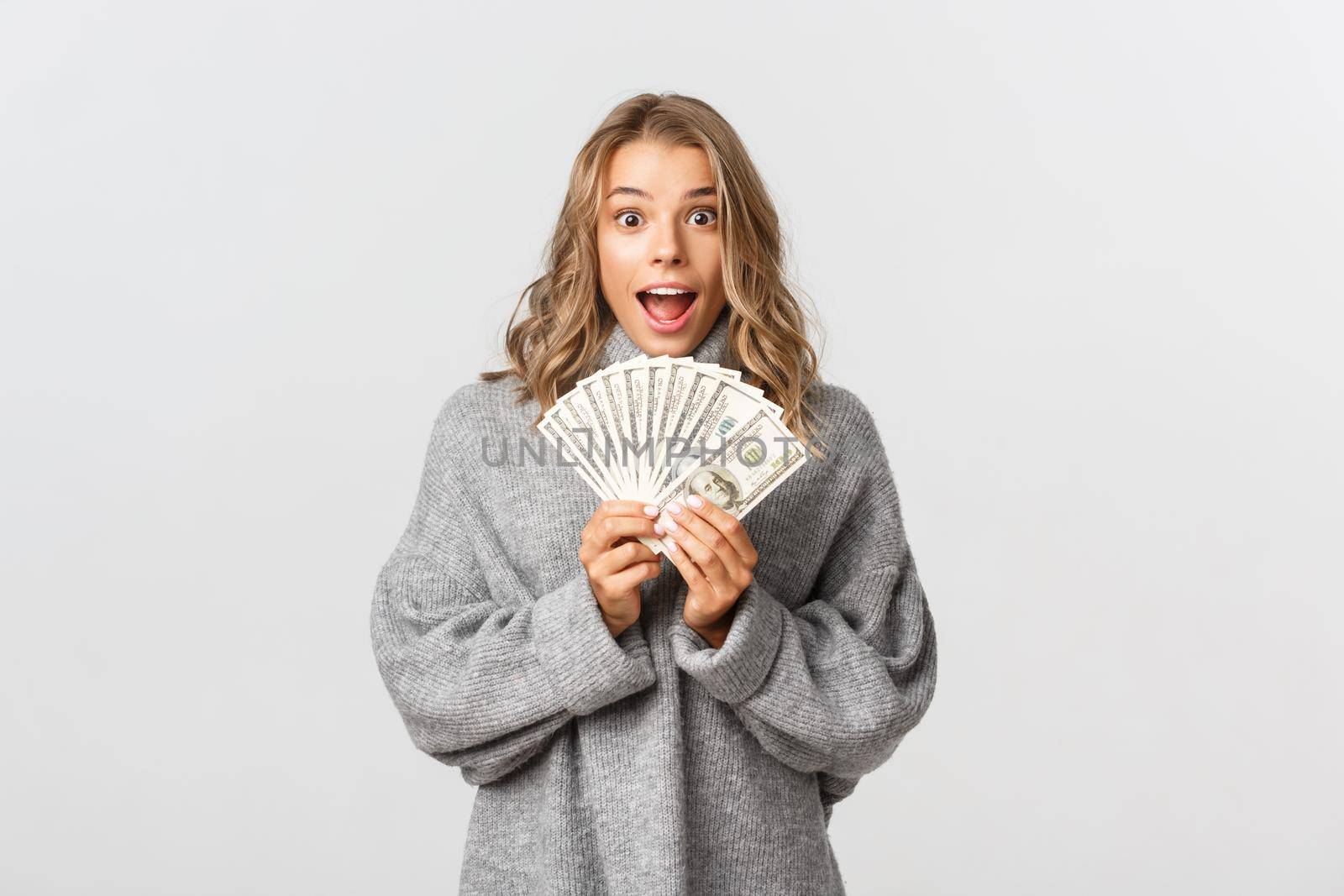 Image of excited attractive girl in grey sweater, holding money and looking amazed, standing over white background by Benzoix