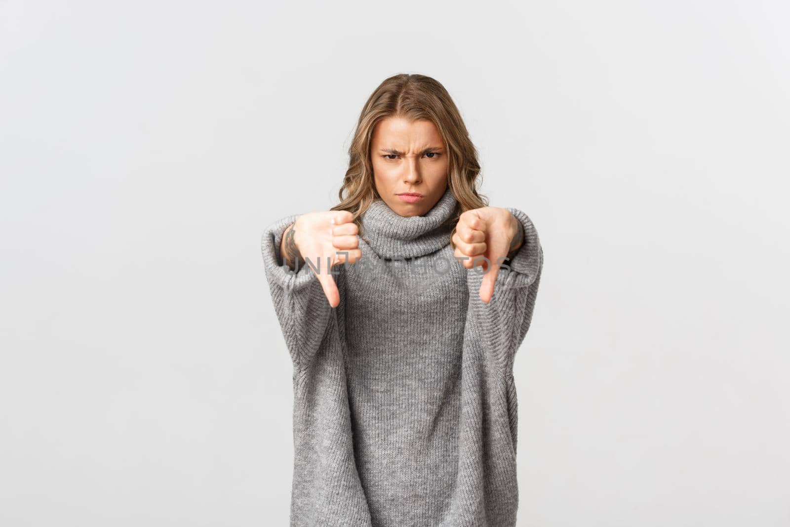 Image of disappointed blond woman in grey sweater frowning upset, showing thumbs-down, judging something bad, standing over white background.