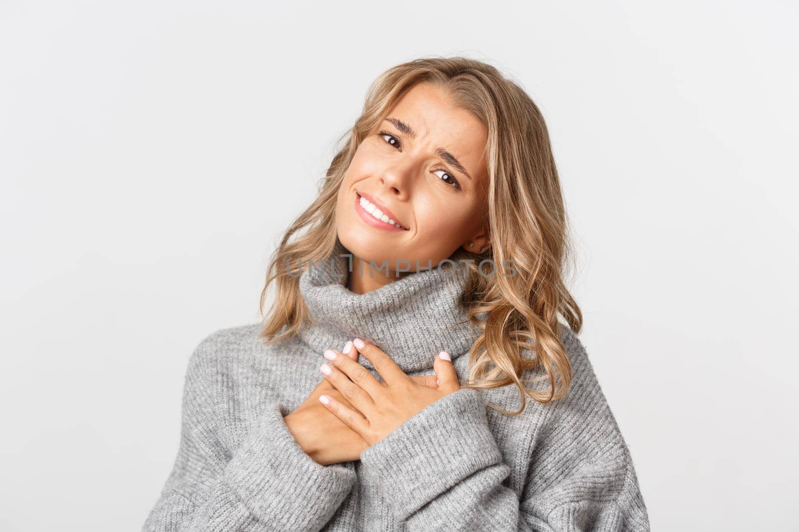 Close-up of beautiful blond girl, looking touched and grateful, holding hands on heart and sighing, standing over white background.