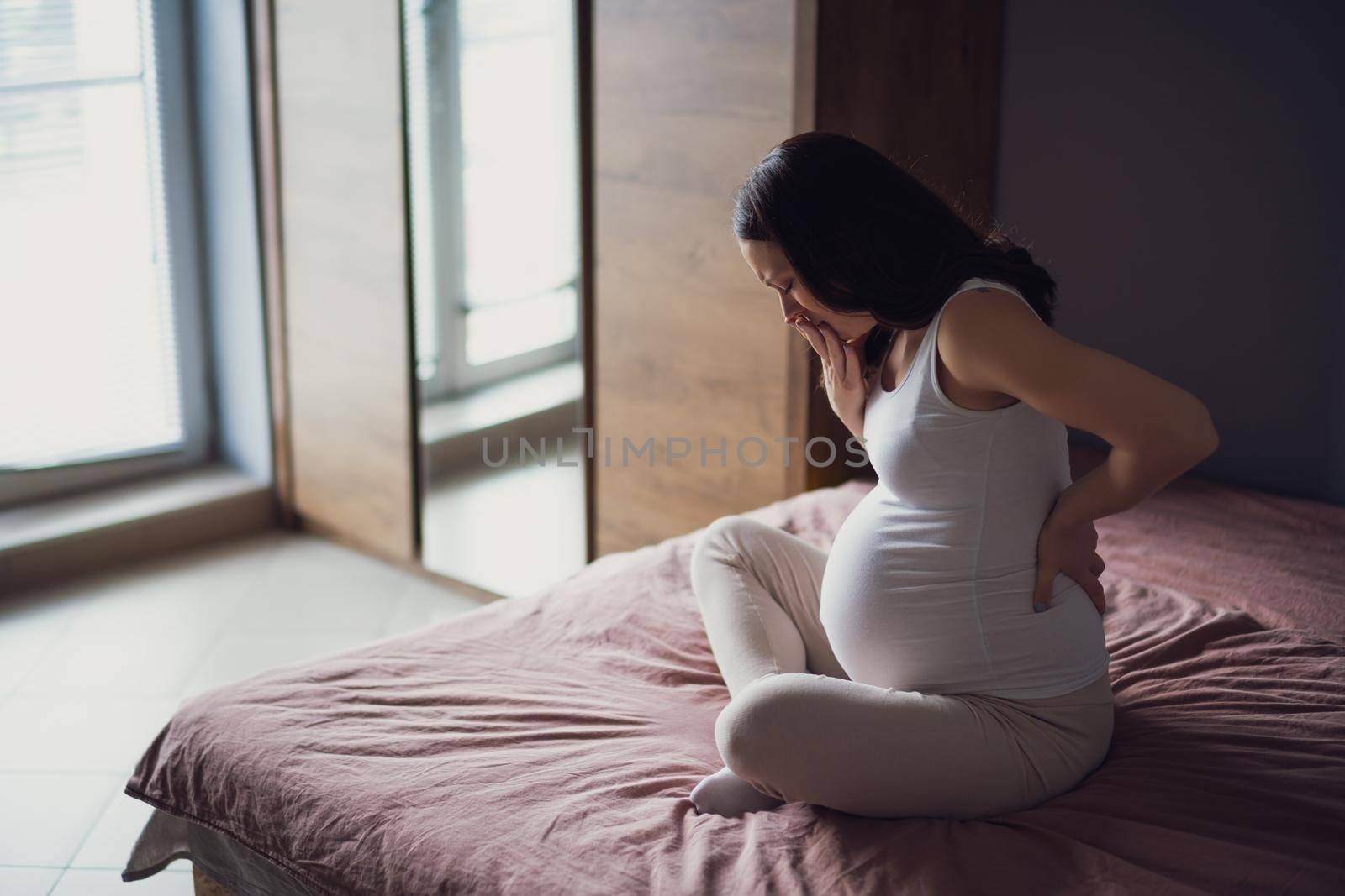 Pregnant woman is having pain in her stomach and back. She is sitting on bed at home.