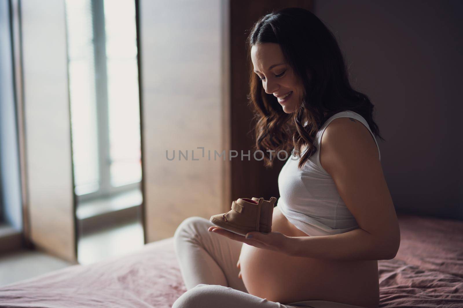 Pregnant woman relaxing at home. She is sitting on bed and holding baby shoes.