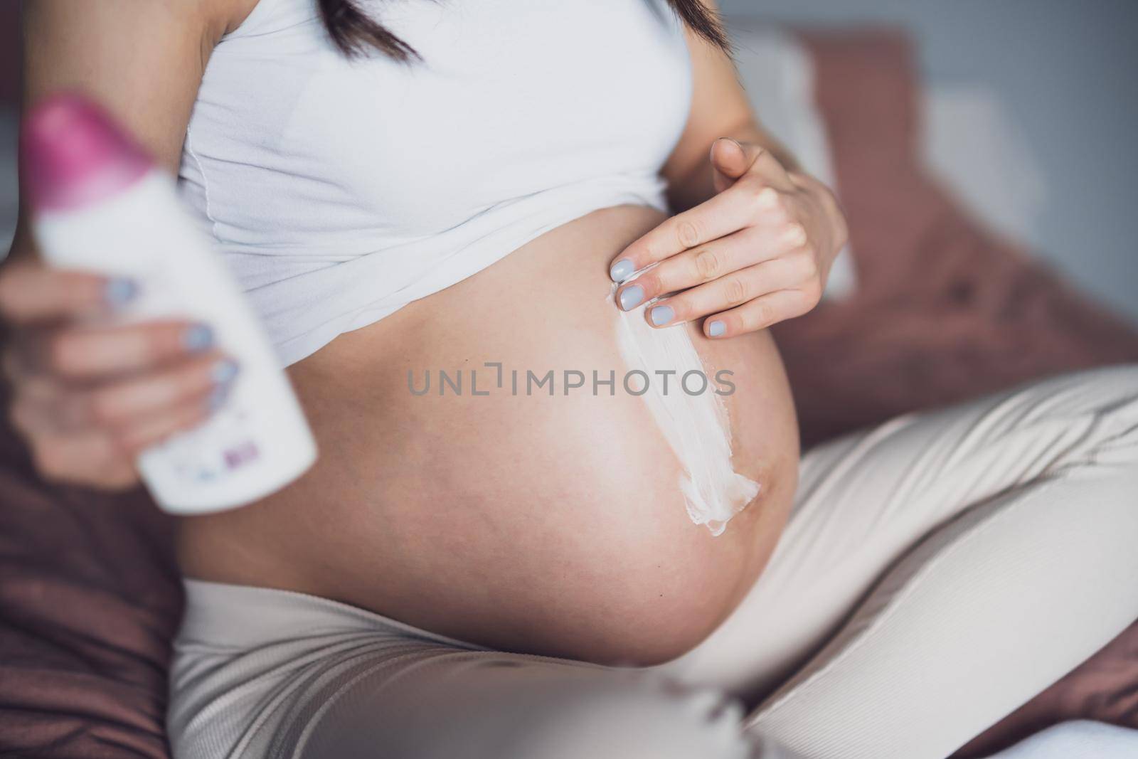 Pregnant woman relaxing at home. She is sitting on bed and applying skin cream to her belly.