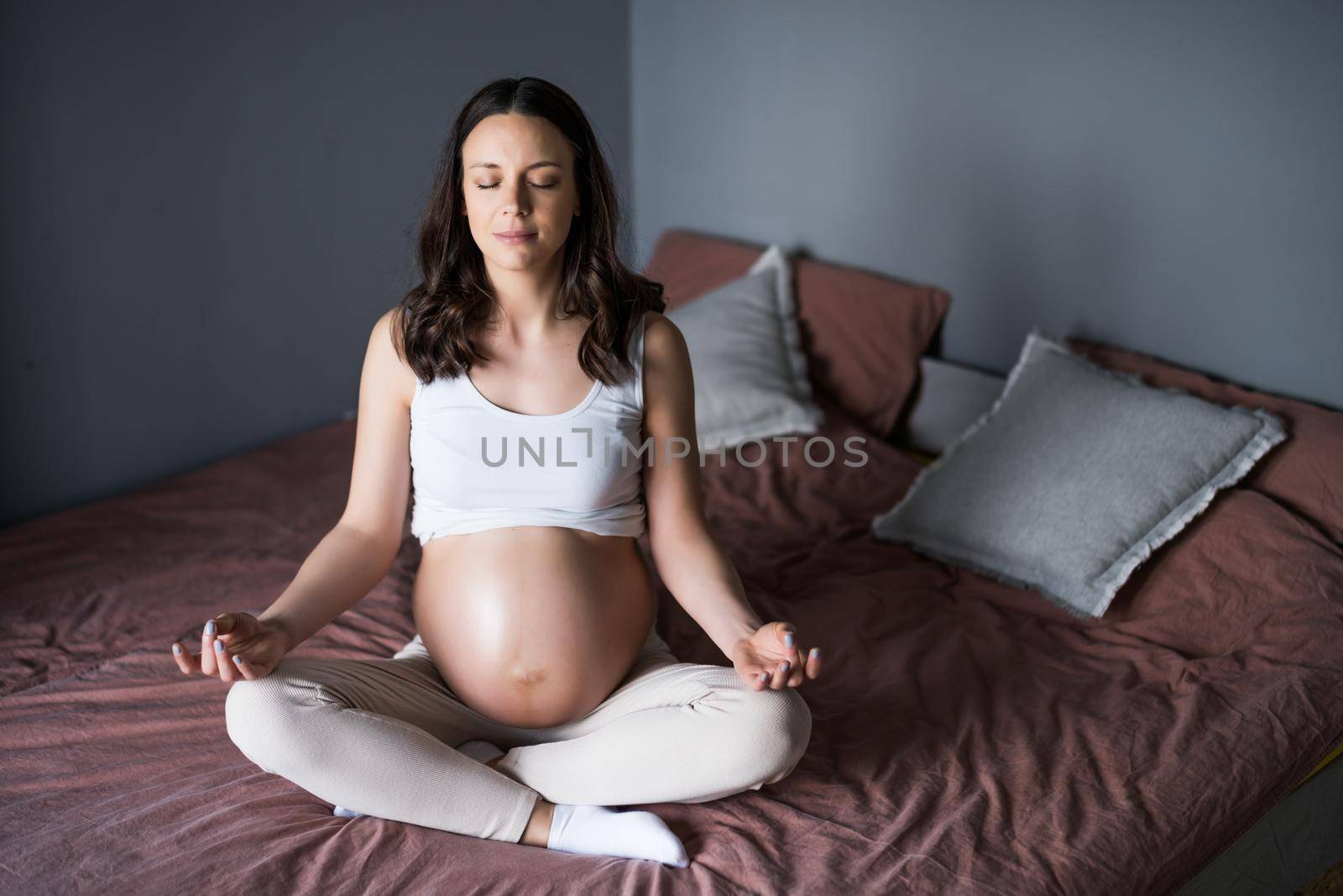 Pregnant woman relaxing at home. She is sitting on bed and meditating.