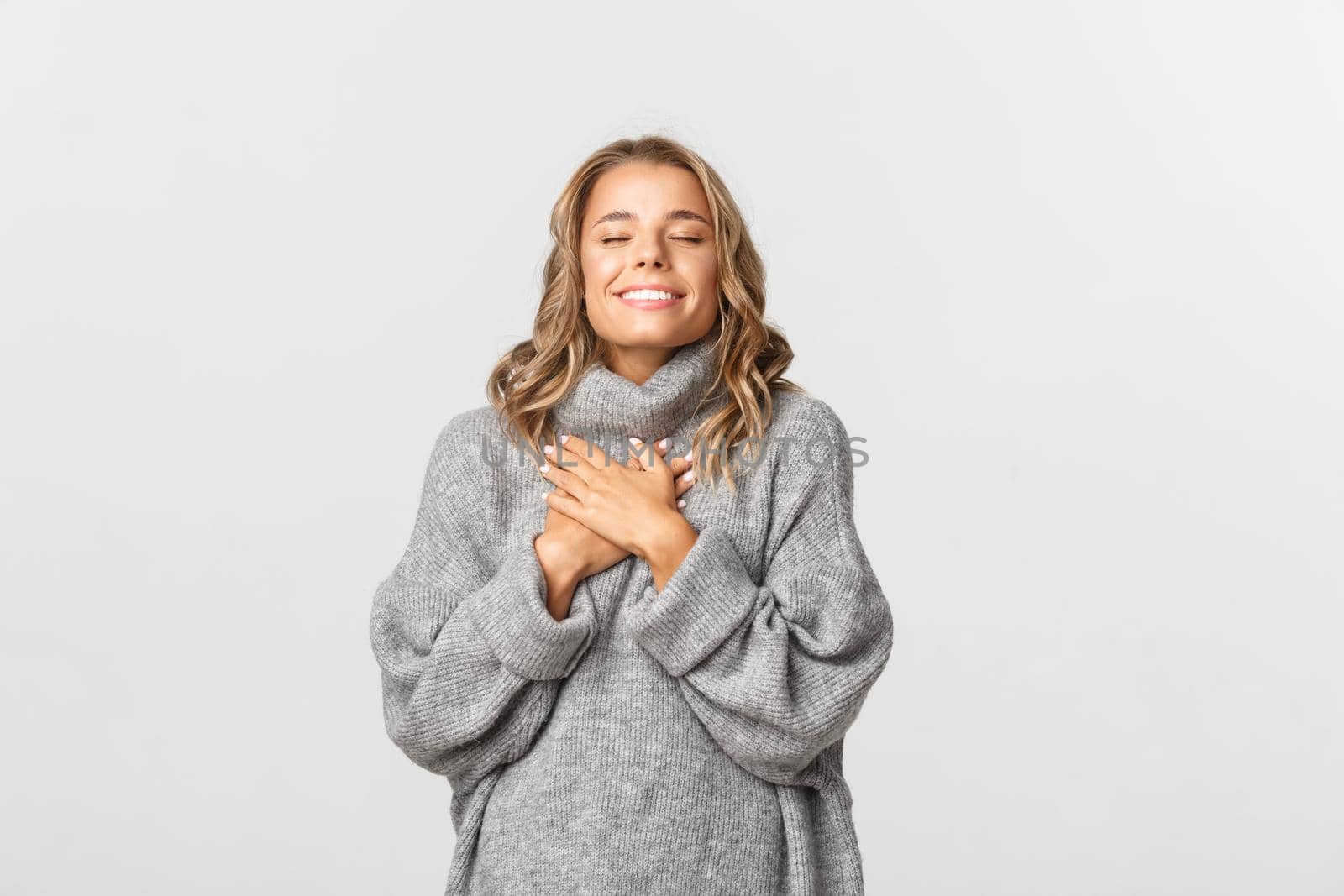 Close-up of beautiful blond girl, wearing grey sweater, feeling nostalgic, holding hands on heart and close eyes, daydreaming over white background.