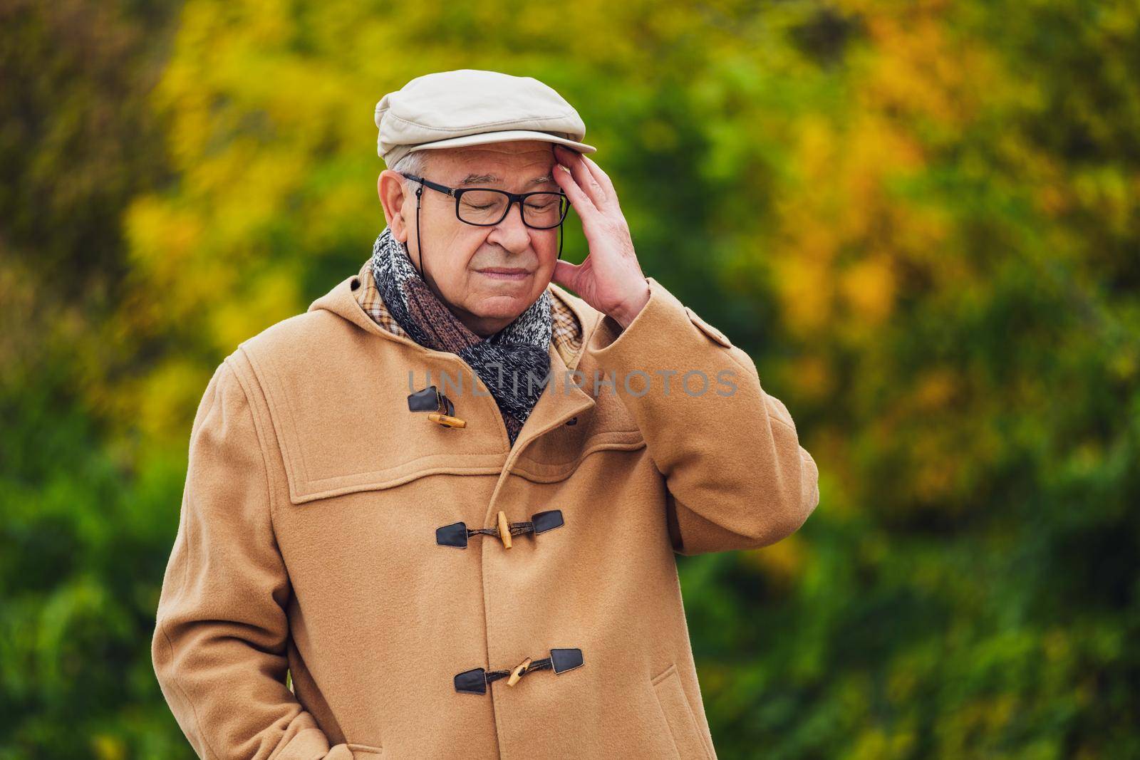 Outdoor portrait of worried senior man in winter coat. He is having headache.