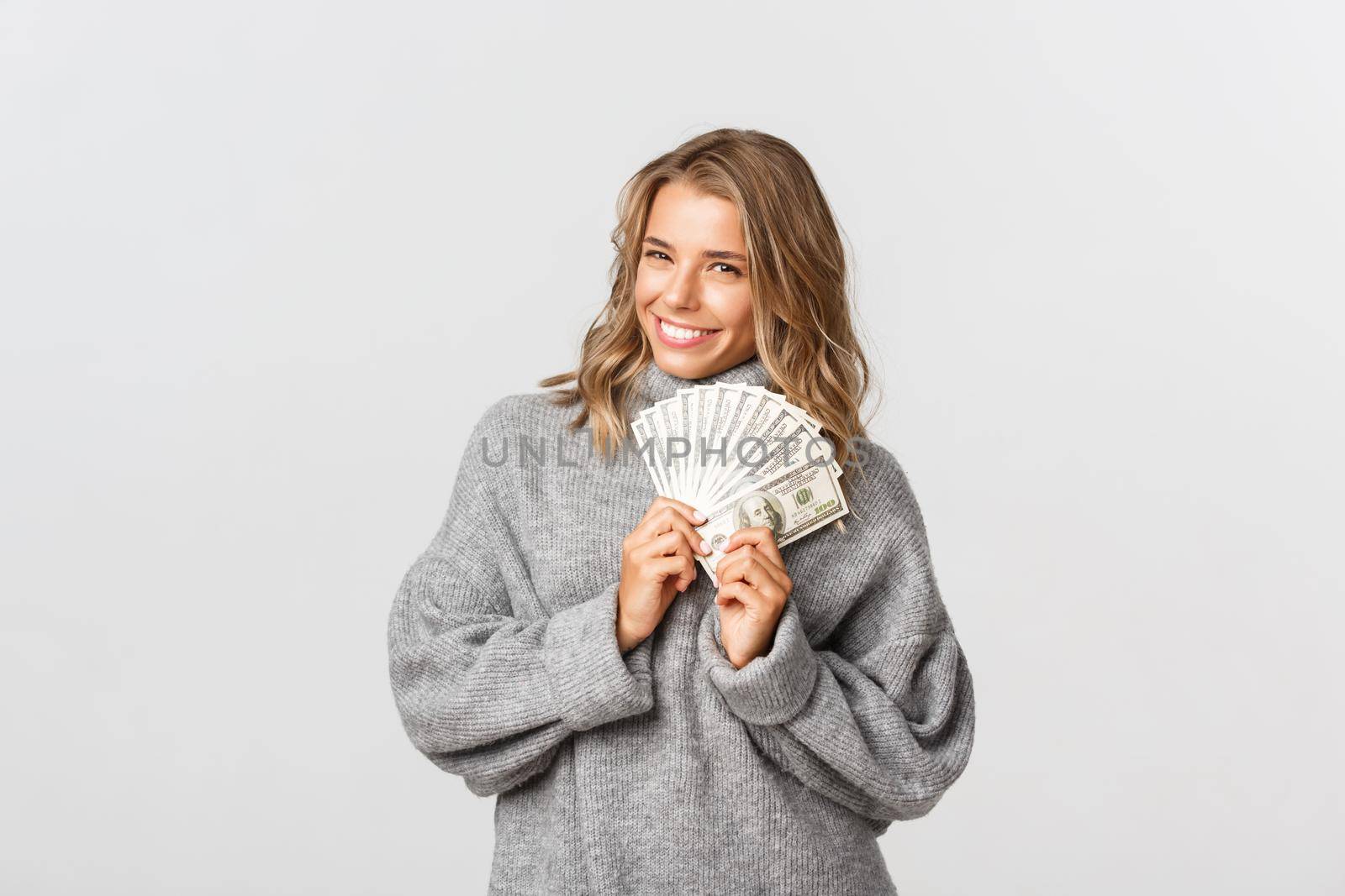 Image of beautiful blond girl in grey sweater, holding money and smiling, thinking how to waste cash, white background.