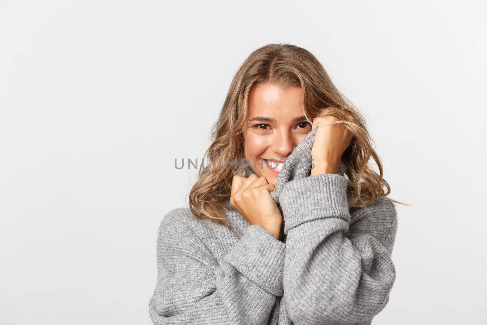Close-up of beautiful romantic girl, touching soft grey sweater and smiling, standing over white background.