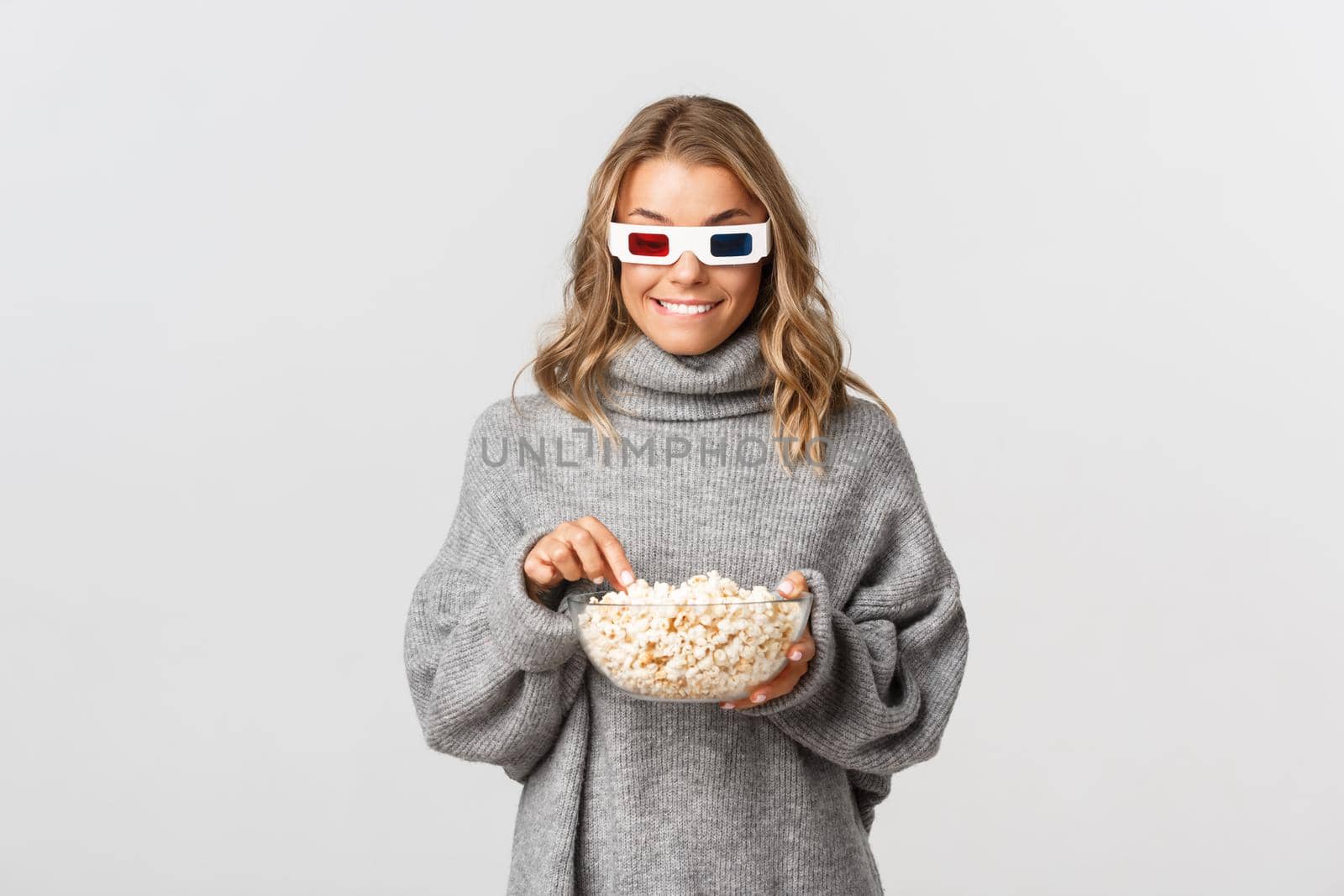 Young pretty girl with blond short hairstyle, wearing 3d glasses, watching movie and eating popcorn, standing over white background.