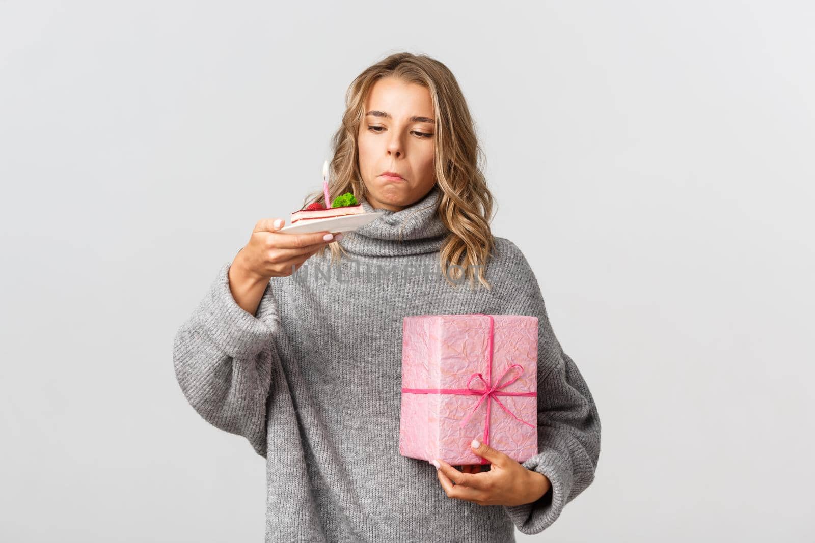 Studio shot of cute blond girl holding gift, looking curious at birthday cake with lit candle, standing over white background.