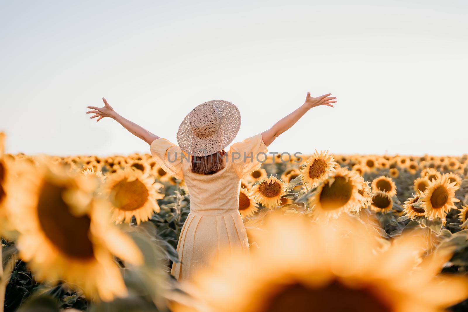 Woman with open arms in sunflowers field. Yellow colors, warm toning. Free girl in straw hat and retro dress. Vintage timeless fashion, amazing adventure, countryside, freedom scene.High quality photo