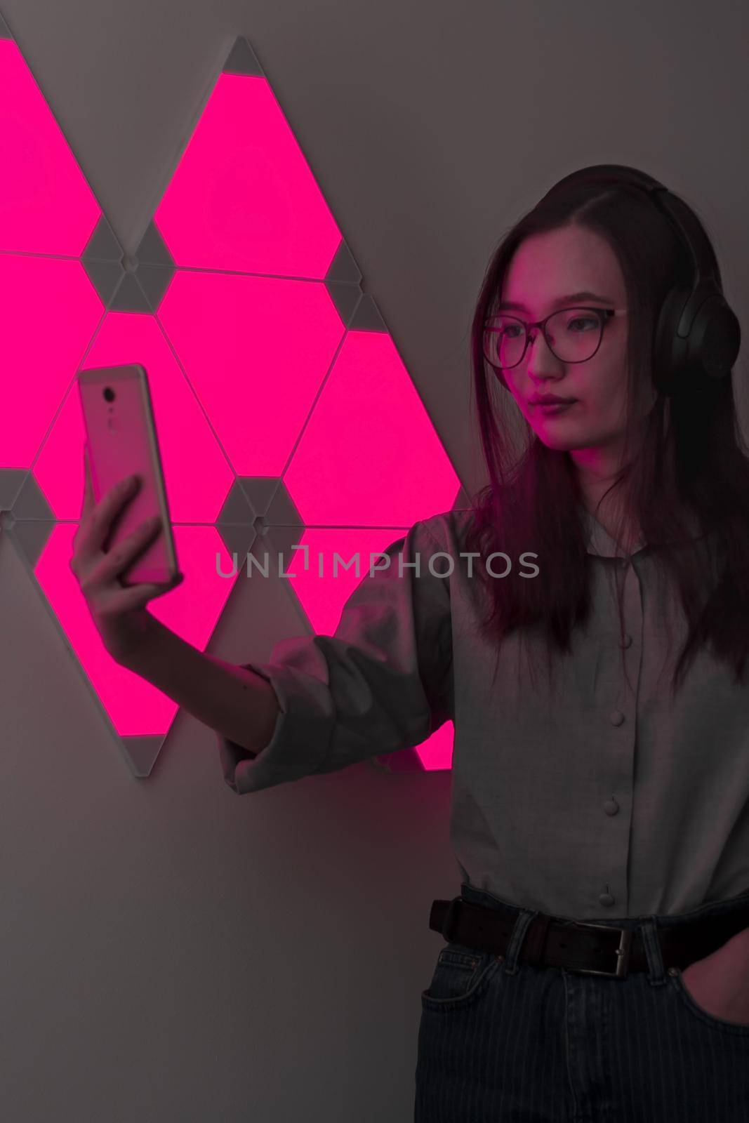 Young pretty woman with wireless headphones in red light of neon panels takes selfie on phone, nightclub and party concept, futuristic neon trend of modern electronic music, dark vertical image