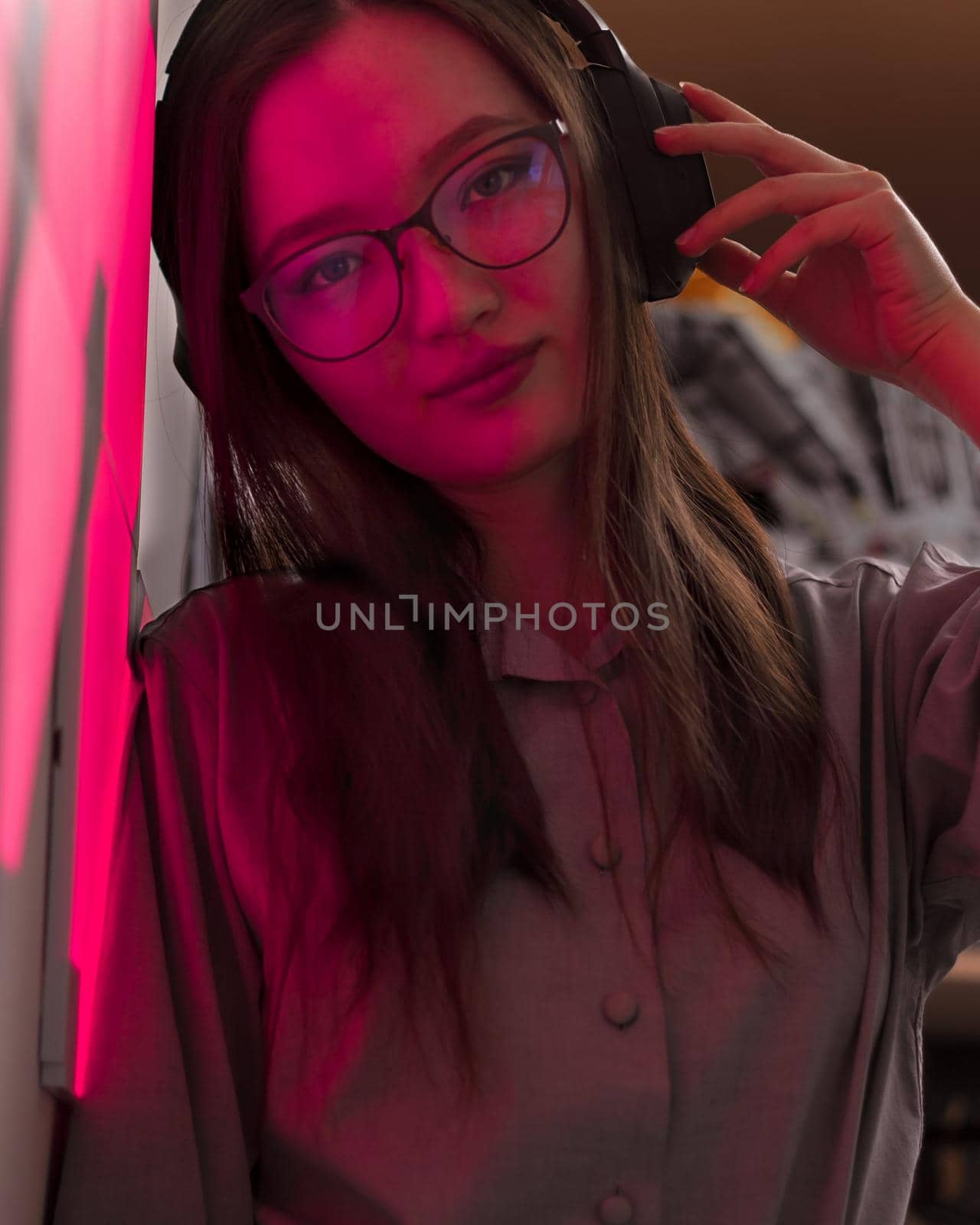 Young smiling woman with wireless headphones in red pink light of neon panels leans on wall looking at lens, nightclub concept, futuristic neon trend of modern electronic music, dark vertical image