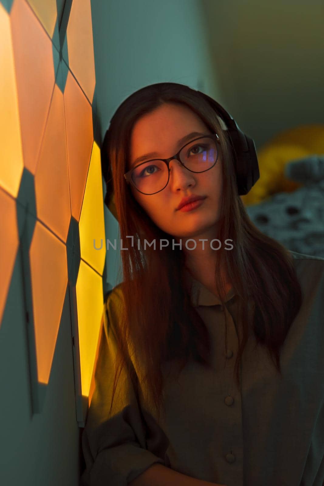 Young pretty brooding woman with wireless headphones in yellow orange light of neon panels listening to music, nightclub concept, futuristic neon trend of modern electronic sound, vertical image