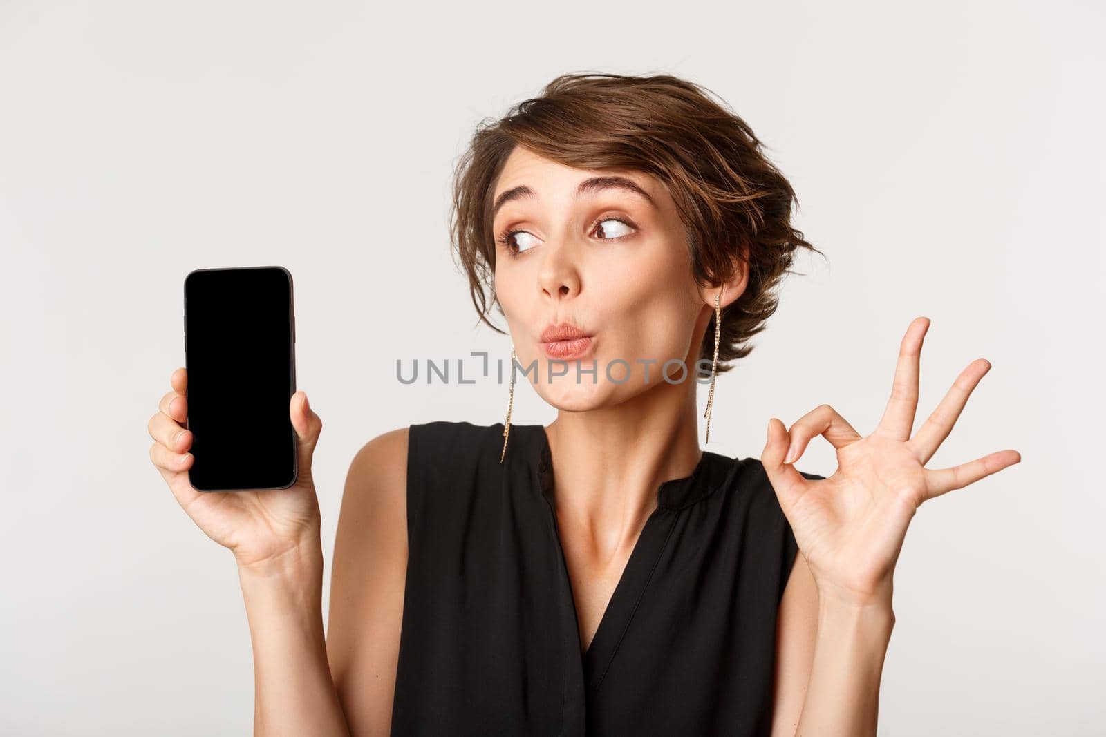 Close-up of silly pretty woman pouting, showing mobile phone screen and okay gesture, standing over white background.