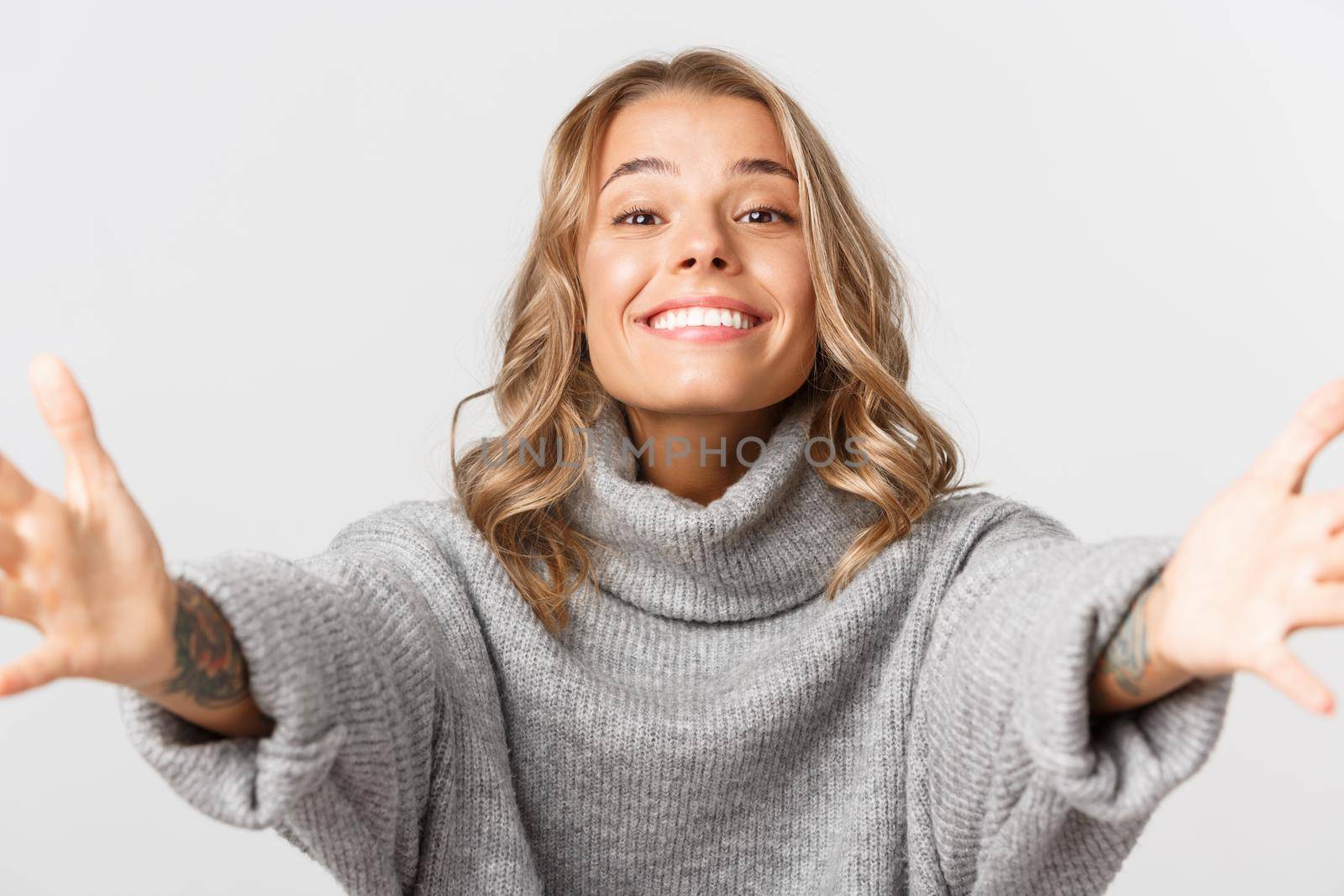 Close-up of cute blond girl in grey sweater, stretching hands forward for hug, receiving something, standing over white background by Benzoix