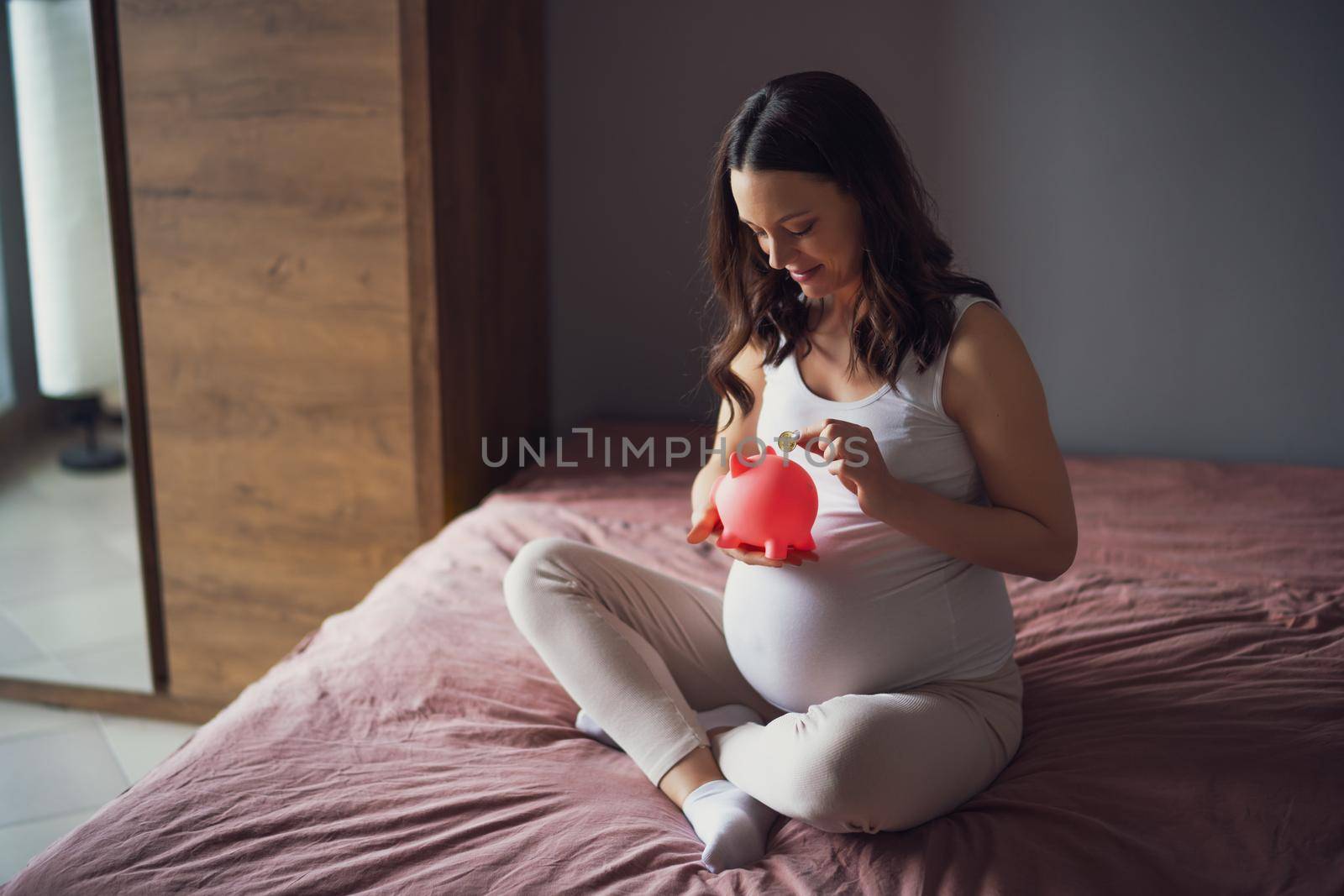 Happy pregnant woman relaxing at home. She is sitting on bed and saving coin in piggy bank.