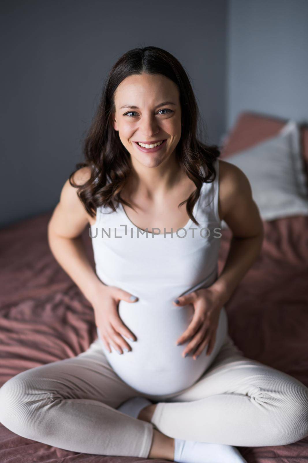 Happy pregnant woman relaxing at home. She is sitting on bed in bedroom.