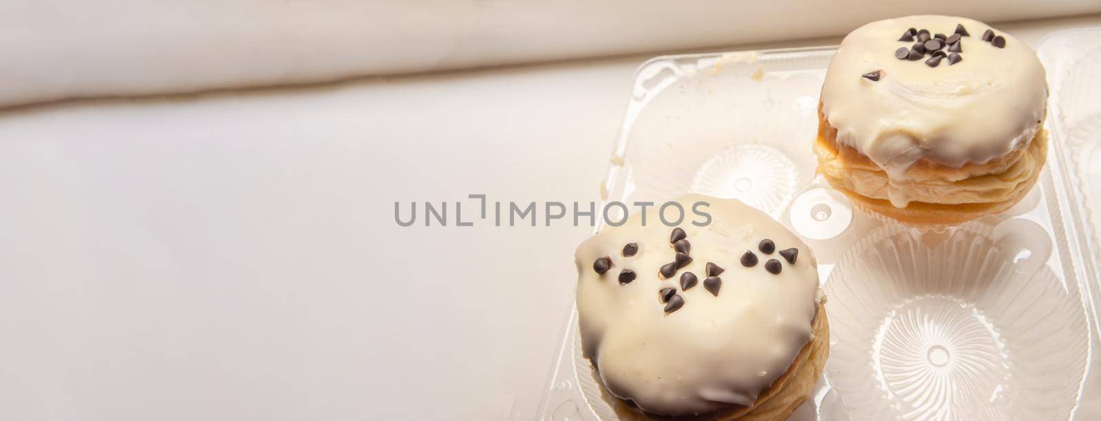 Two delicious donuts with icing sugar and chocolate chips on a white background in a plastic transparent container, a banner with a copy of the space on the left.