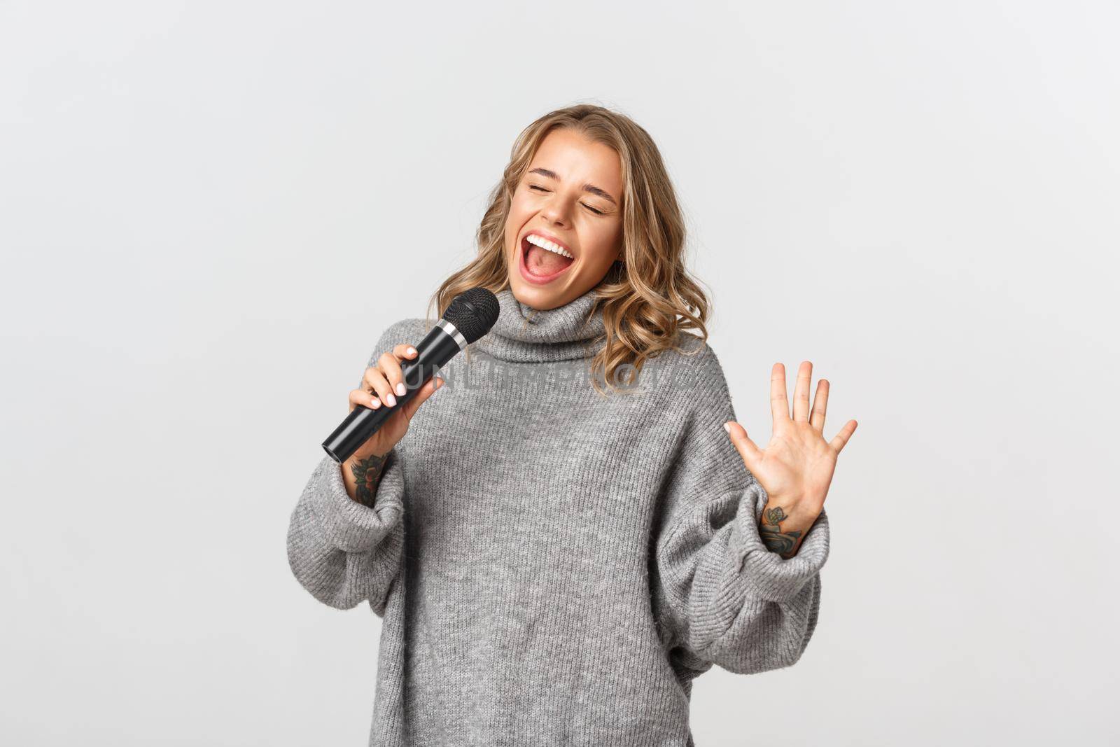 Image of happy blond girl in grey sweater, singing with microphone, standing over white background.