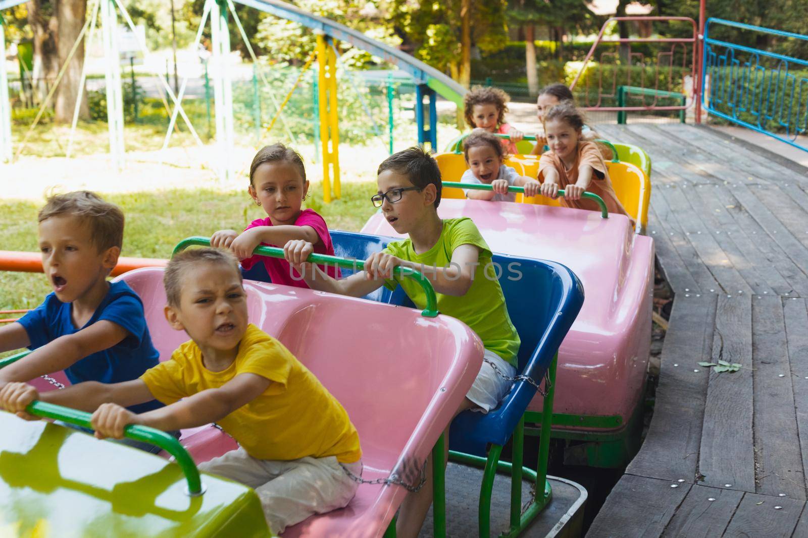 The group of happy little children are riding a roller coaster in the park. The children are dressed in a colorful summer clothes