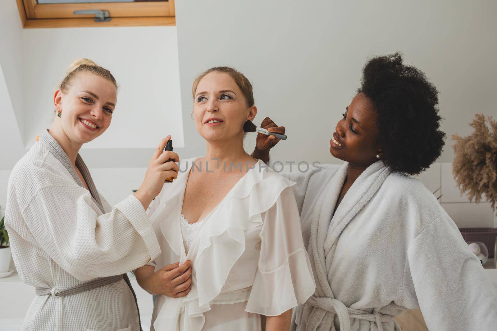 Three smiling young women in bathrobes have fun at the spa and beautify each other