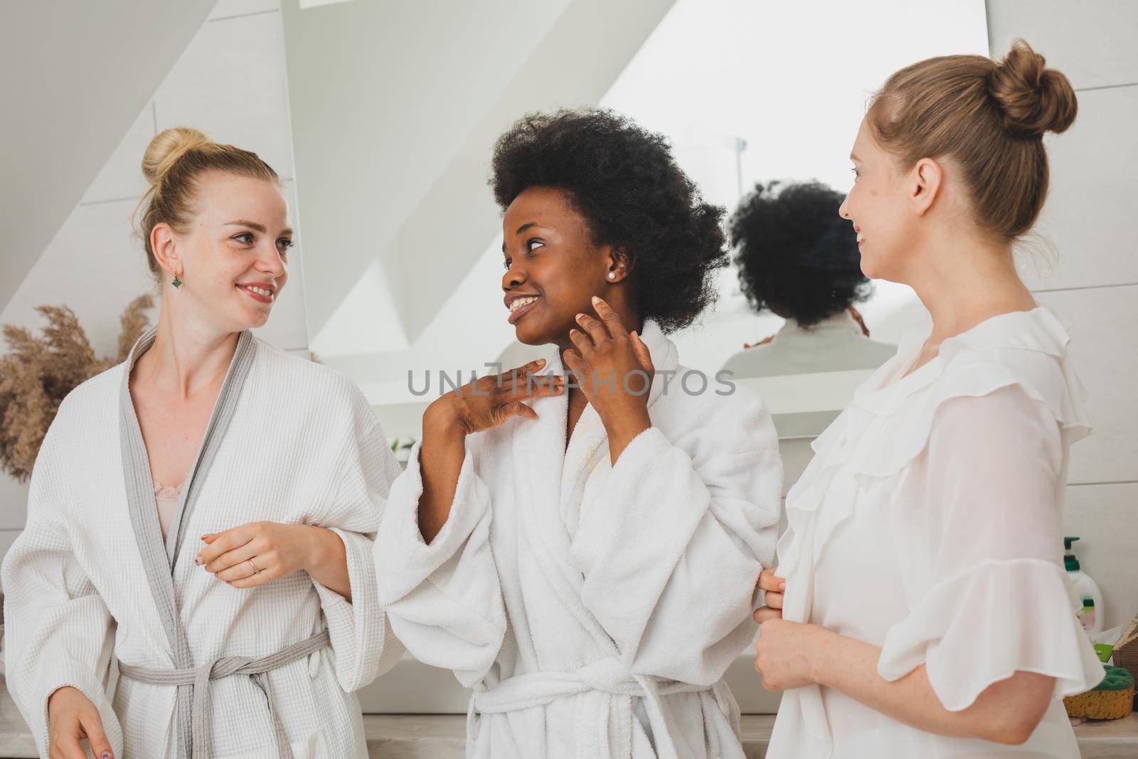 Portrait of three happy young women in bathrobes. They hold facial cleansing tools and look at the camera