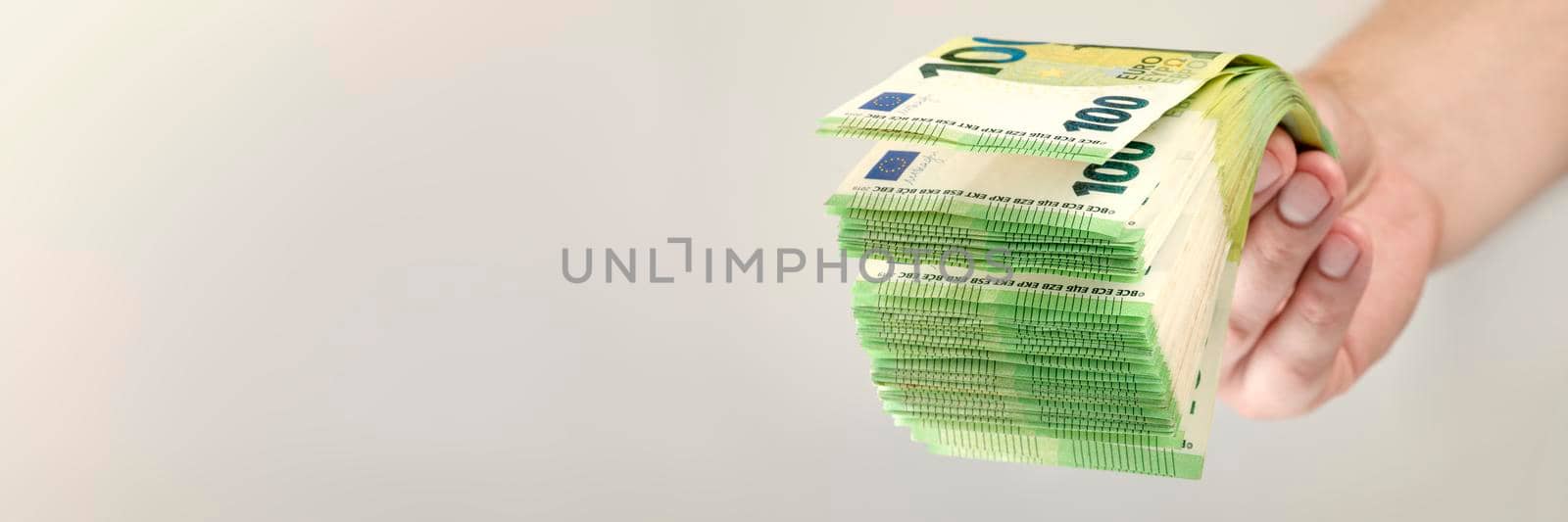 Stack of euro bills in hand. A large stack of 100 euro banknotes in a male hand on a uniform gray background. The concept of financial assistance, real estate purchase, loan or insurance payment. by SERSOL