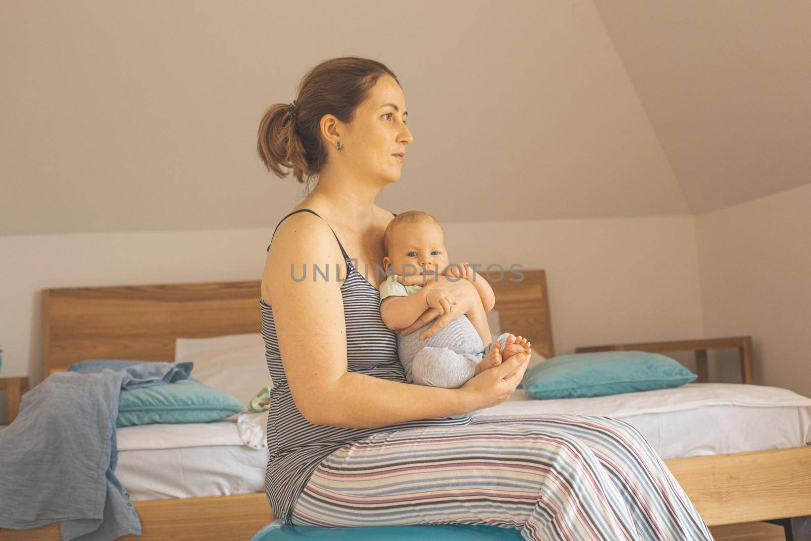 Mother holding and rocking baby sitting on fitball. New lifestyle with asleep newborn at bedroom