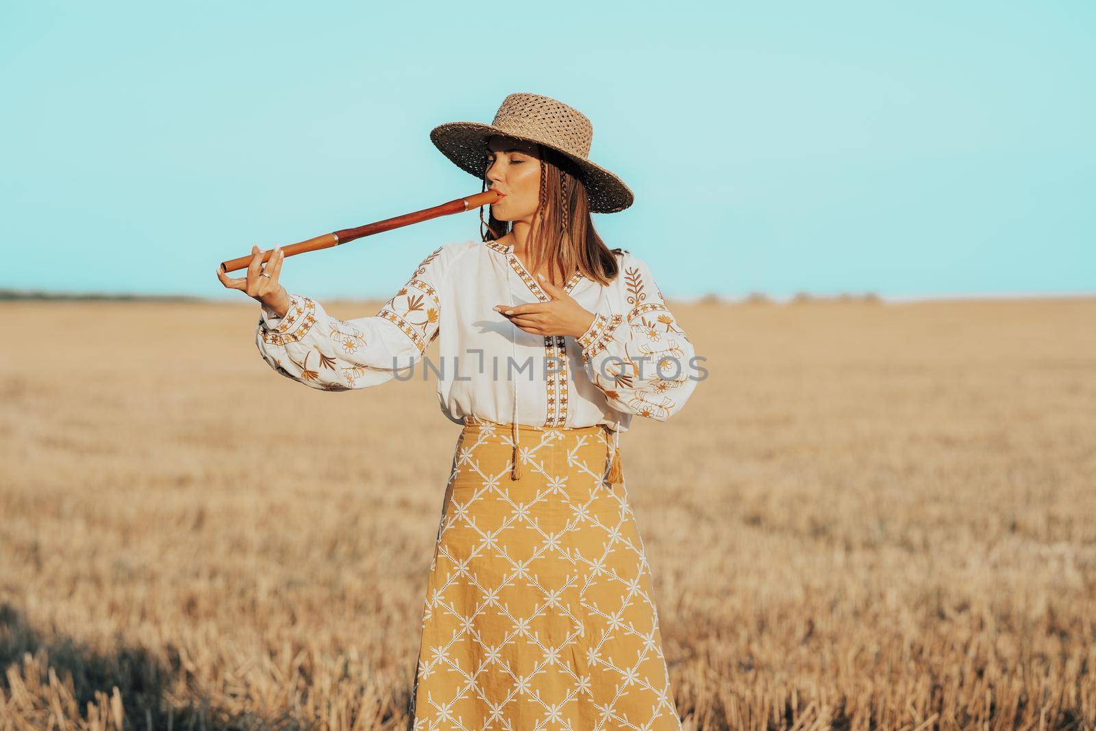 Woman playing on woodwind wooden flute - ukrainian telenka or tylynka in wheat field. Folk music concept. Musical instrument. Musician in traditional embroidered shirt - Vyshyvanka. High quality photo