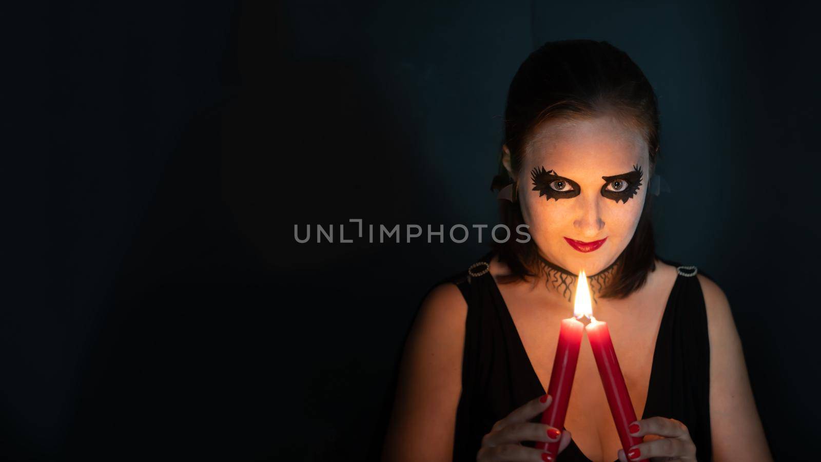 Girl in image of witch looks straight into frame with burning candle in hands on black background. Concept of Halloween and magic. Copy space by Laguna781