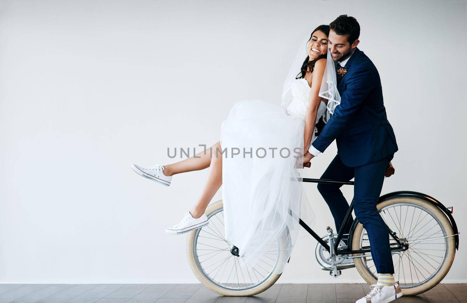 Off to our honeymoon. Studio shot of a newly married young couple riding a bicycle together against a gray background