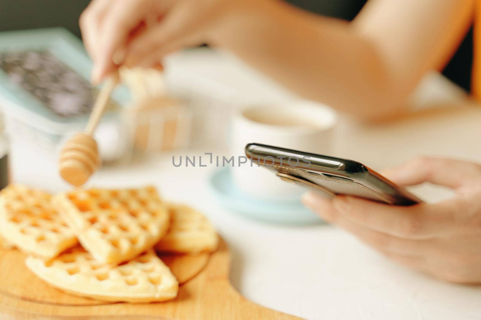 Morning romantic pastime. Online communication over lunch. Woman writes messages on cell phone and breakfast. Belgian waffles on desk, cup of coffee and honey stick on white table.